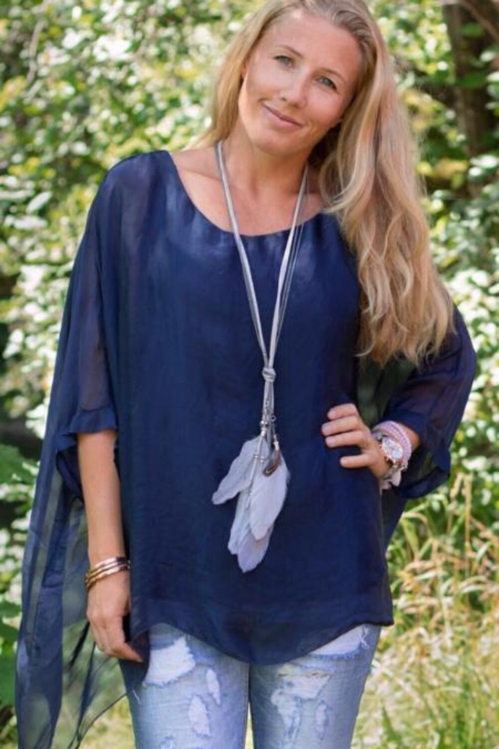Blond kvinna står utomhus i grönskande park och ler mot kameran, med handen på höften. Hon bär en marinblå, luftig tunika med slitsärm, slitna ljusblå jeans och ett långt halsband med fjädrar.