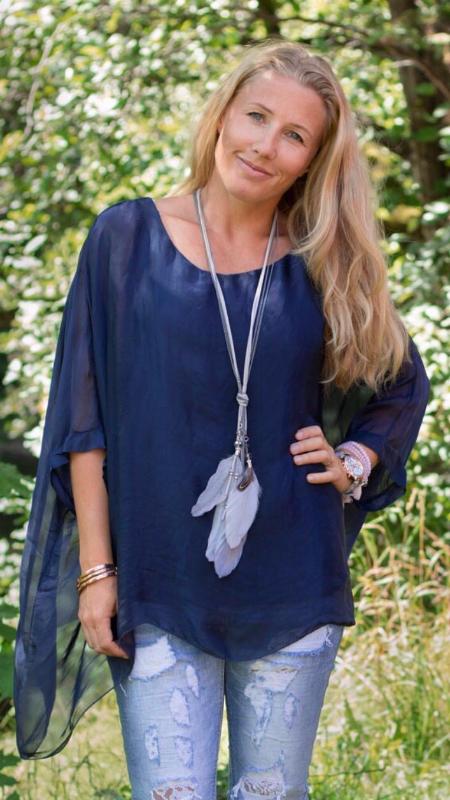 Blond kvinna står utomhus i grönskande park och ler mot kameran, med handen på höften. Hon bär en marinblå, luftig tunika med slitsärm, slitna ljusblå jeans och ett långt halsband med fjädrar.
