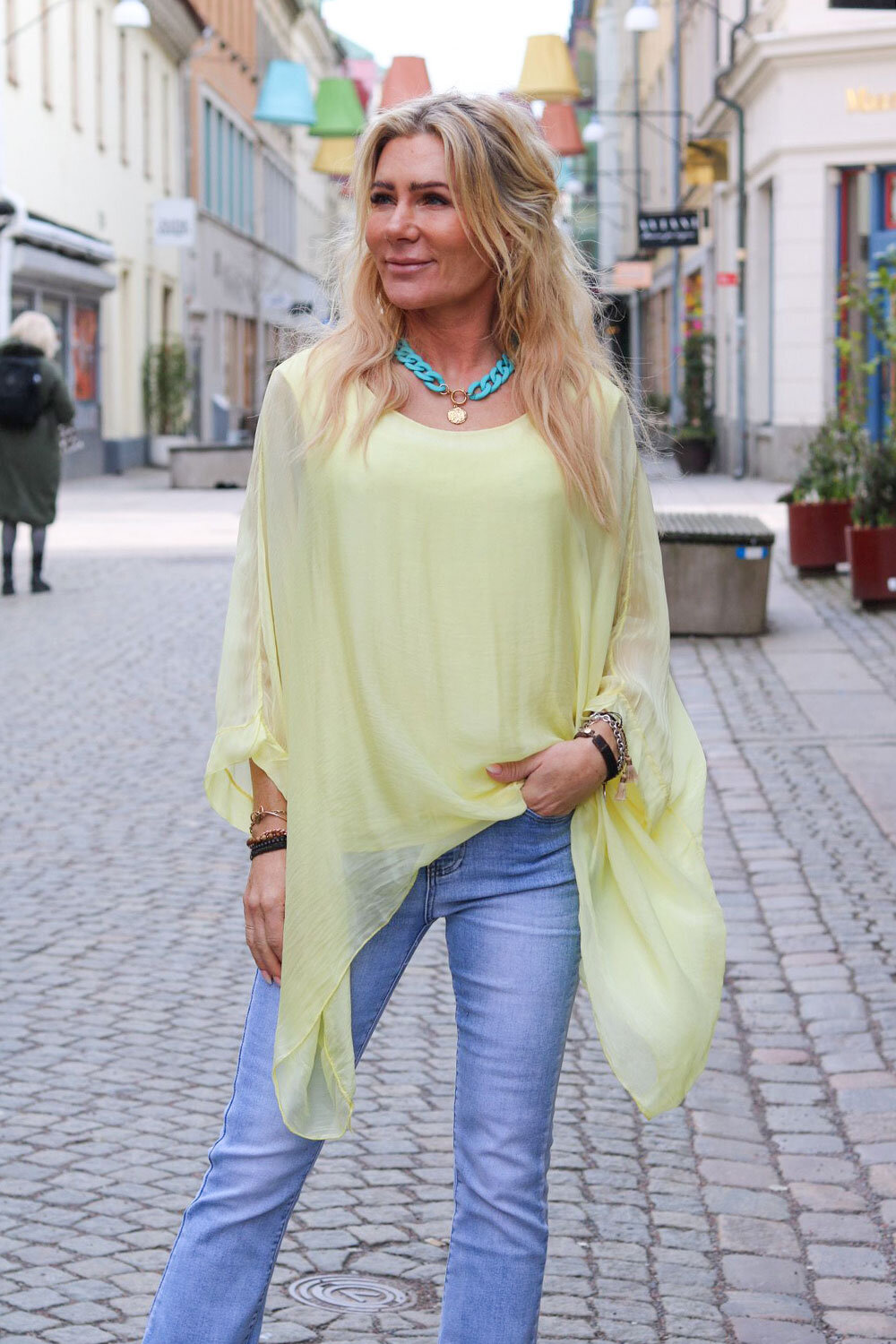 Kvinna står på en kullerstensgata i stadsmiljö och tittar åt sidan, klädd i en enfärgad ljusgul, luftig silkestunika med vida ärmar över jeans. Bär turkost halsband och armband; färgglada lampskärmar hänger ovanför.