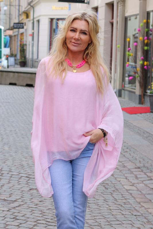 Kvinna med långt blont hår promenerar på en kullerstensgata och tittar mot kameran, klädd i enfärgad ljusrosa, luftig silkestunika över jeans, med rosa halsband och armband; butiksfasader syns i bakgrunden.