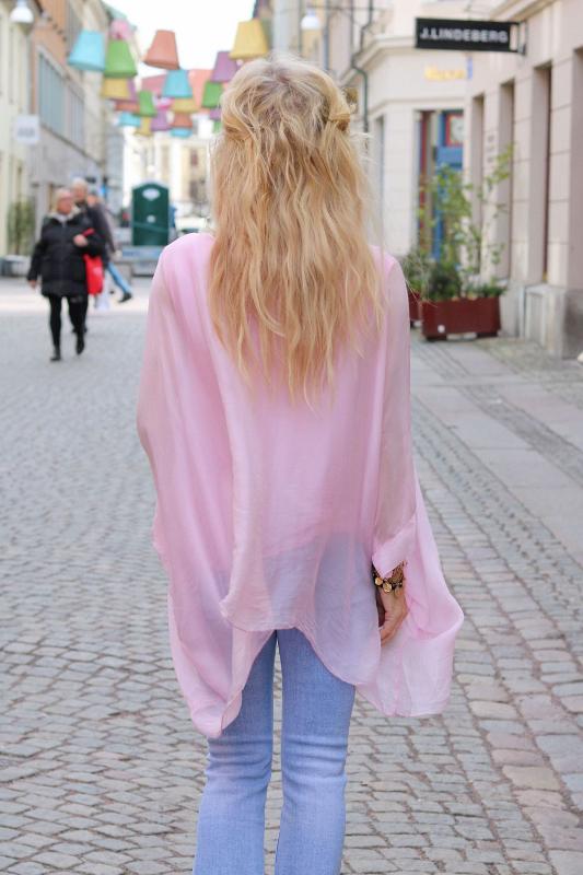 Kvinna sedd bakifrån promenerar på en kullerstensgata, klädd i en ljusrosa, enfärgad silkestunika med vid, draperad passform över ljusblå jeans. Färgglada hängande lampor syns ovanför och butiker längs gatan.