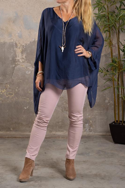 Kvinna står i studio och visar en enfärgad navyblå silkestunika med vid, flödande passform och långa ärmar. Hon bär ljusrosa smala byxor, beige ankelboots, armband och långt halsband mot rå betongvägg.