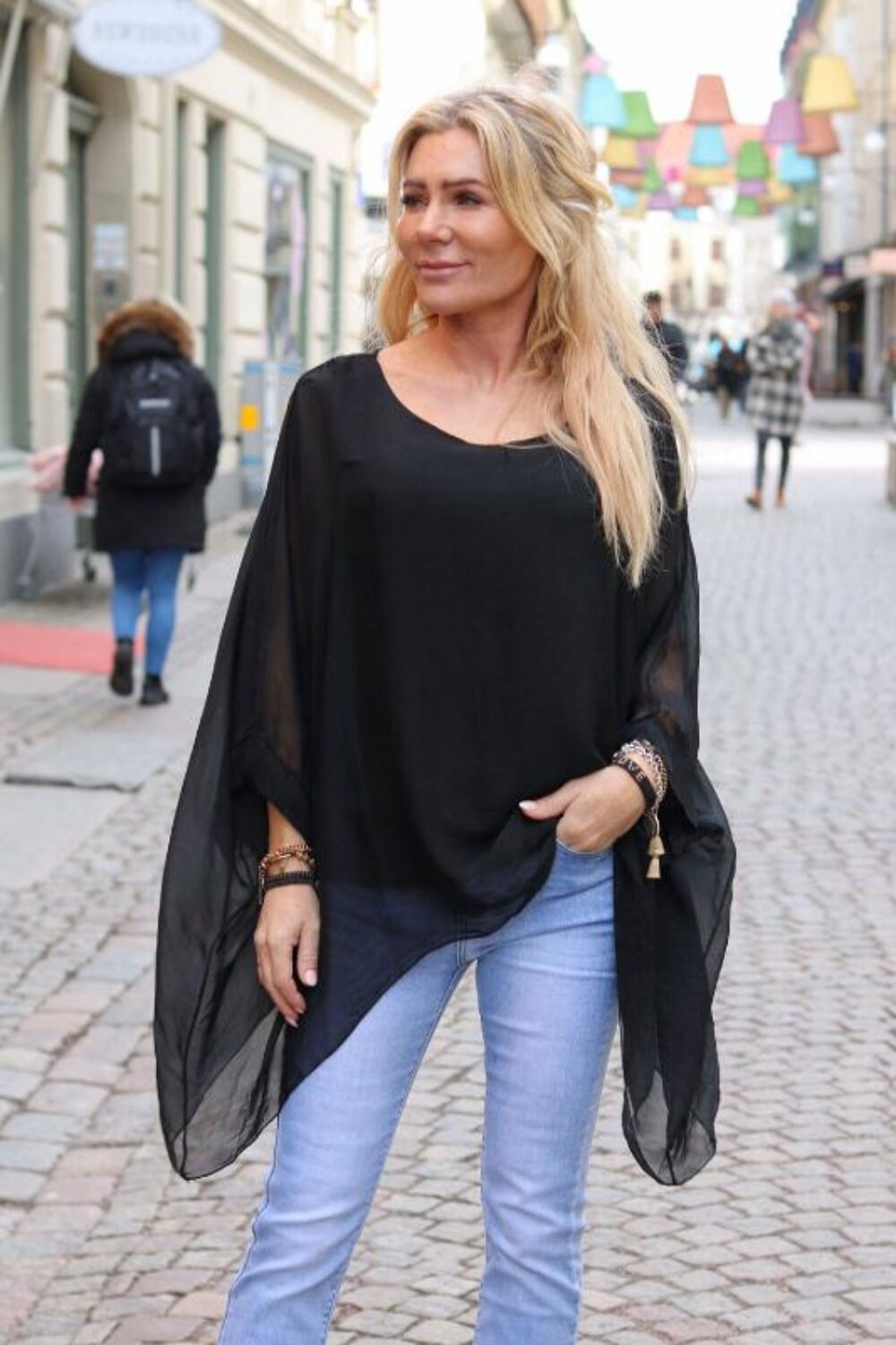 Kvinna står på en kullerstensgata och poserar i en svart enfärgad, luftig silkestunika med vida, transparenta ärmar. Hon har ljusblont hår, bär ljusblå jeans och armband; färgglada lampor och fotgängare syns i bakgrunden.