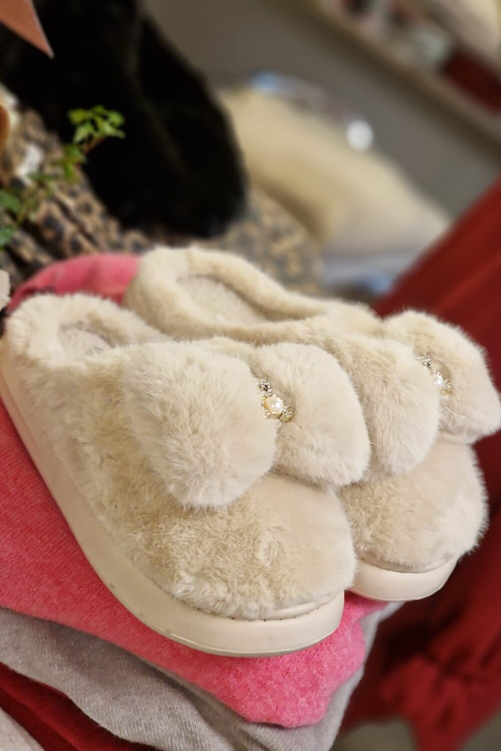 Ett par beige, fluffiga tofflor ligger ovanpå vikta rosa och grå plagg. Tofflorna har stora rosetter med pärldekoration och glittriga stenar, fotograferade i närbild med suddig bakgrund inomhus.