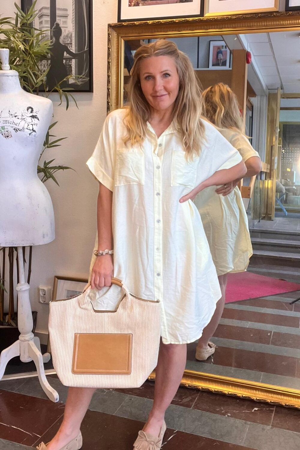 Blond kvinna står och poserar med ena handen på höften och håller en stor beige tygväska. Hon bär en vit skjortklänning och ljusa loafers, framför en guldramsspegel i en butikslik miljö.