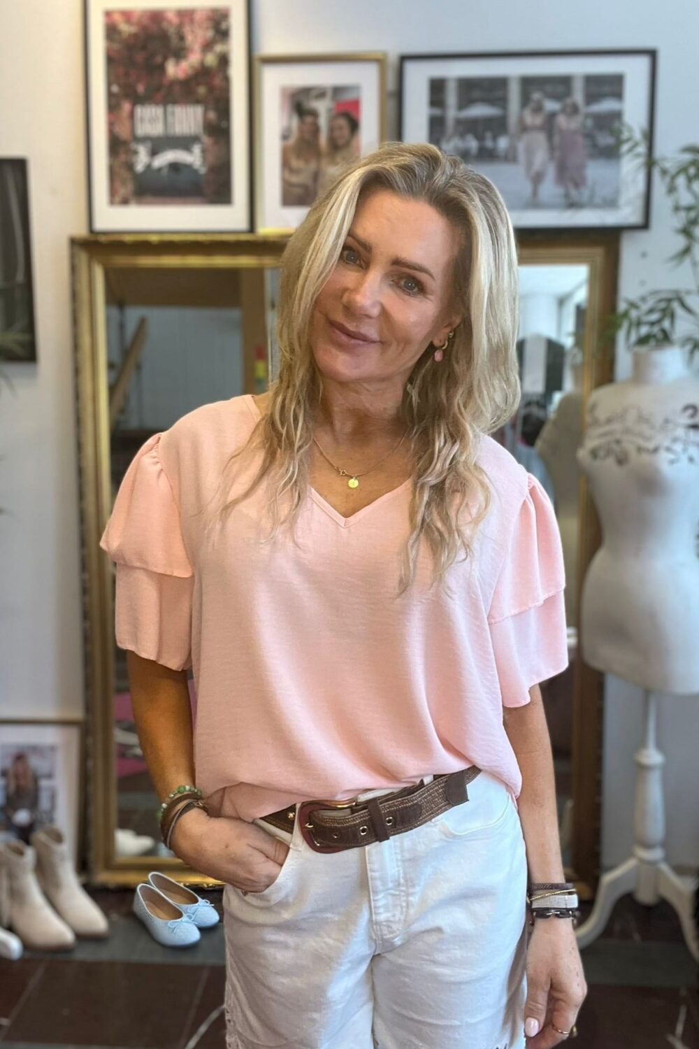 Blond kvinna står och ler mot kameran med ena handen i fickan, klädd i ljusrosa volangtopp och vita shorts med brunt bälte. Bakom syns guldspegel, inramade foton på väggen och skor på golvet.