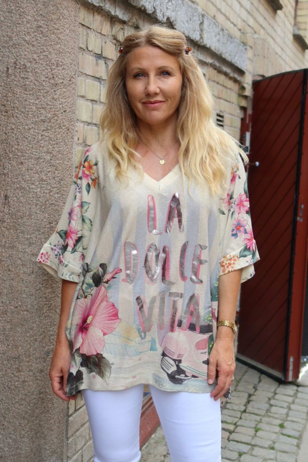 Kvinna med långt blont hår står och ler mot kameran i en smal gränd vid en tegelvägg och röd port. Hon bär vit byxa och blommig tunika med texten “LA DOLCE VITA”.