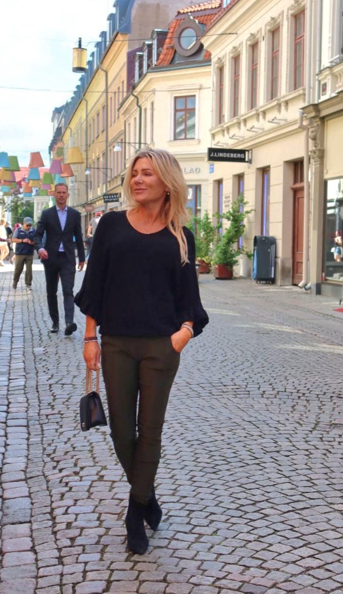 Vaaleahiuksinen nainen kävelee mukulakivikadulla, käsi taskussa ja pieni käsilaukku toisessa kädessä. Taustalla näkyy ohikulkijoita, värikkäitä riippuvaloja ja kivitaloja; kyltti “J.LINDEBERG” erottuu kadun varrella.