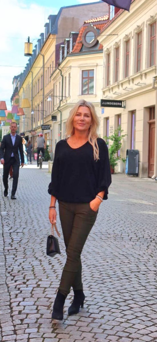 Vaaleahiuksinen nainen kävelee itsevarmasti mukulakivikadulla, käsi taskussa ja pieni käsilaukku toisessa kädessä. Taustalla näkyy värikkäitä katukoristeita, ohikulkijoita ja liikkeitä; kyltissä lukee “J.LINDEBERG”.