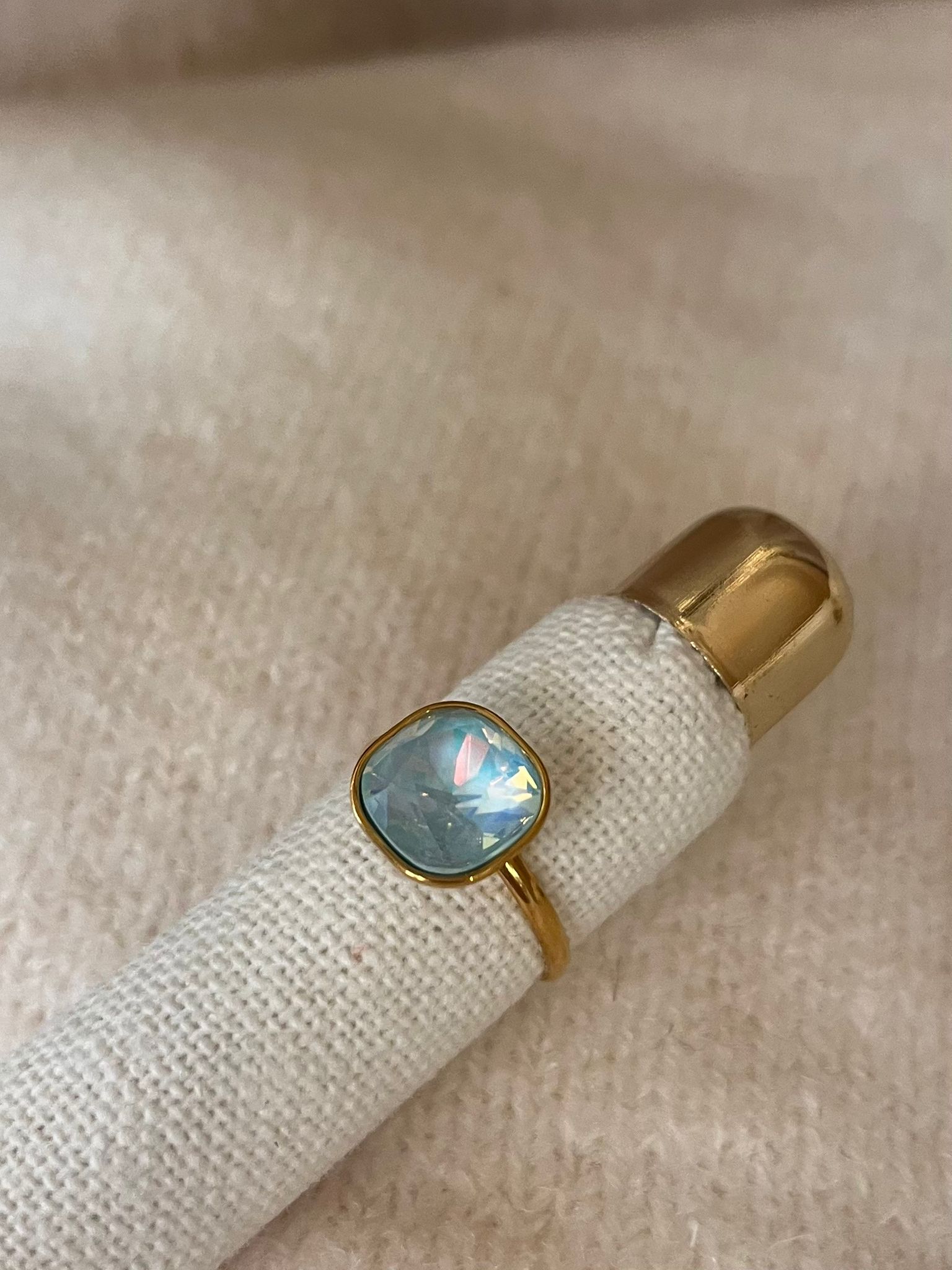 Guldfärgad ring med oval, facetterad ljusblå sten som glittrar i flera färger, ligger runt en vit tygklädd ringhållare med guldtopp. Närbild mot mjuk, beige bakgrund.