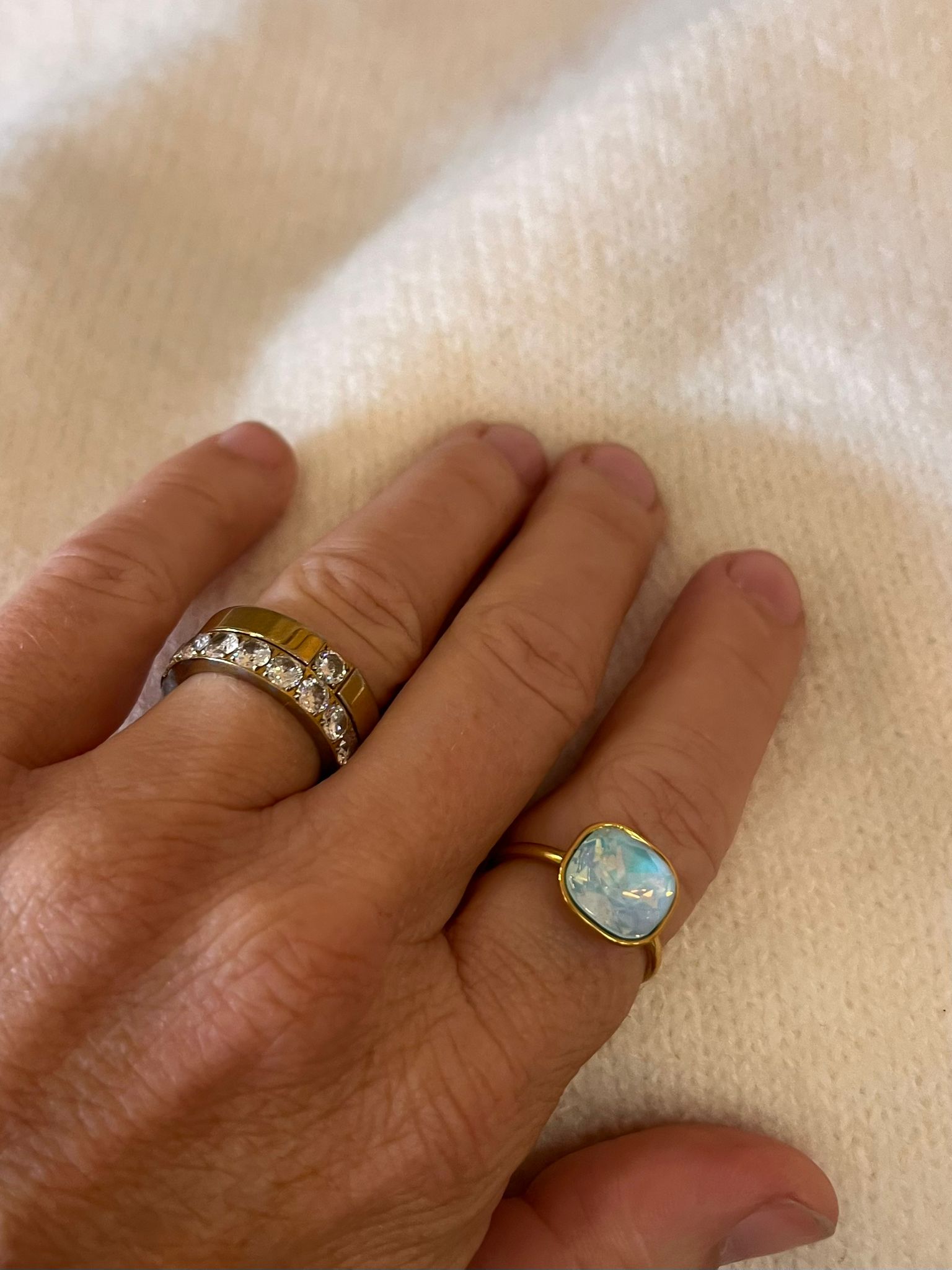 En hand vilar på en ljus, mjuk filt och visar två ringar: en bred guldfärgad ring med infattade klara stenar samt en guldring med stor ljusblå, oval sten. Ingen synlig text.