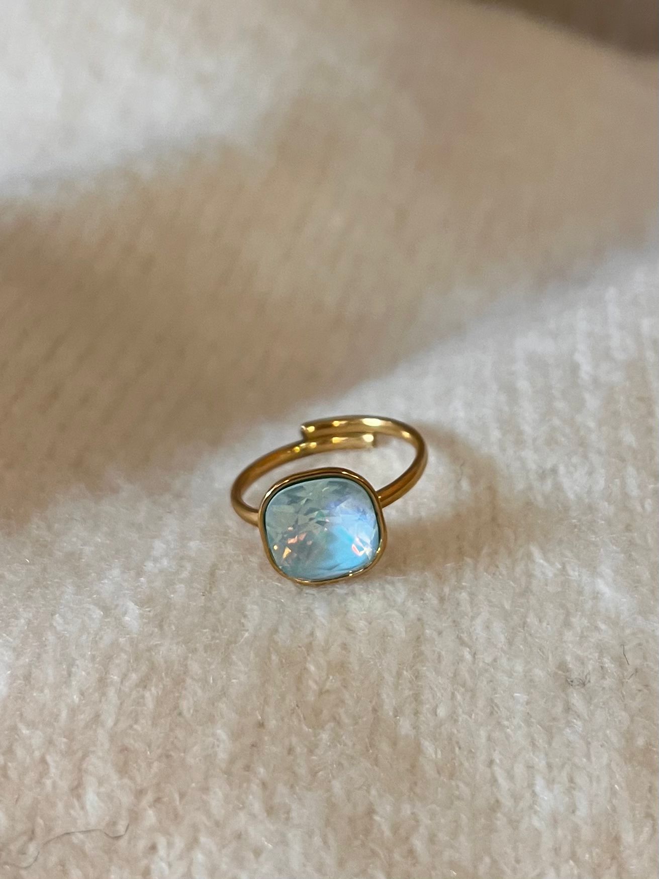 Tunn guldfärgad ring med öppen skena ligger på ett mjukt beige tyg. Den har en rund, ljusblå skimrande sten i infattning, fotograferad nära med fokus på stenens reflektioner och detaljer.