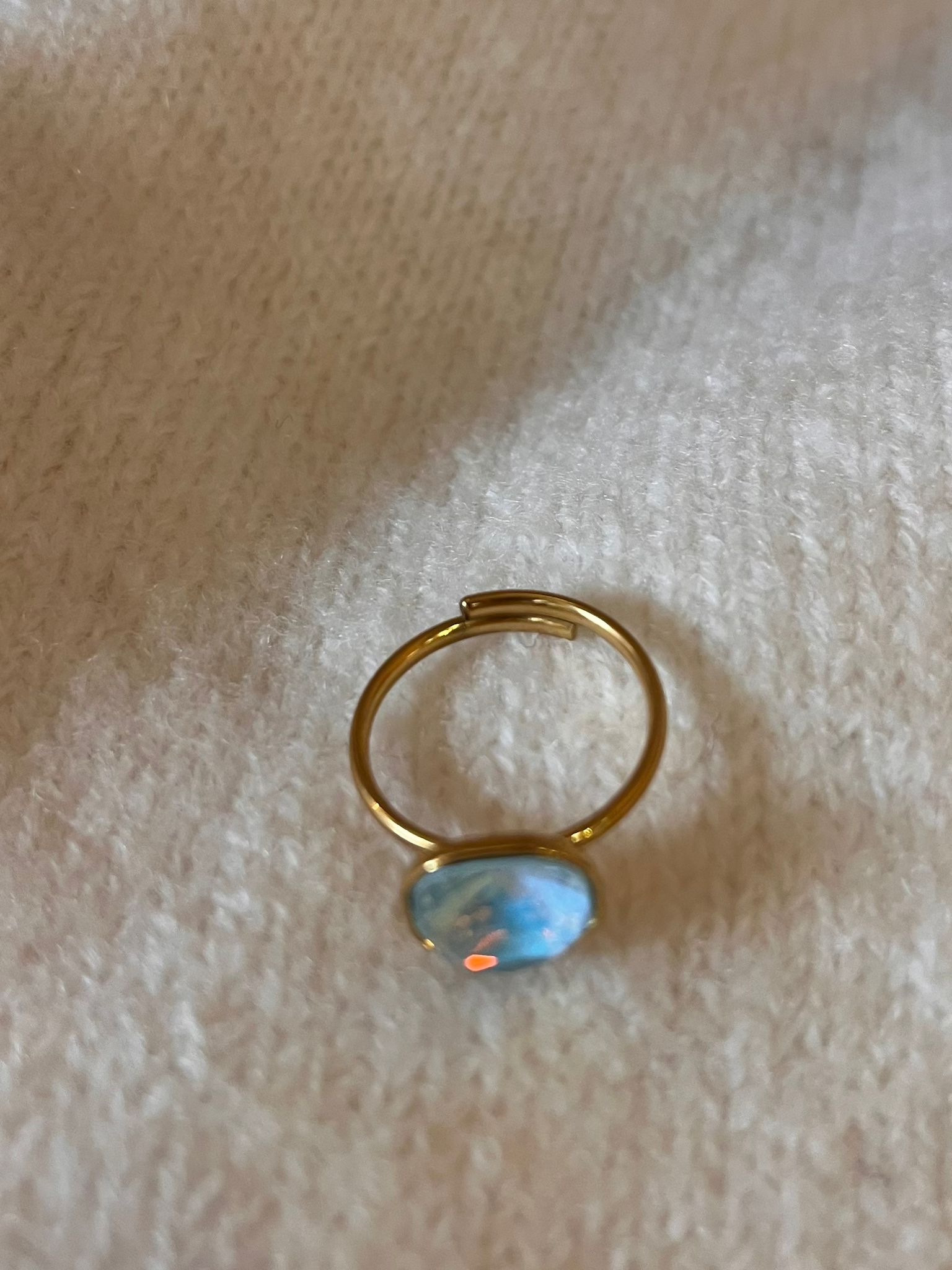 Guldfärgad ring med öppen skena ligger på ett vitt, mjukt tyg. En oval, opal-liknande sten i ljusblått med små orange reflexer sitter i en tunn infattning nära ringens nederkant.