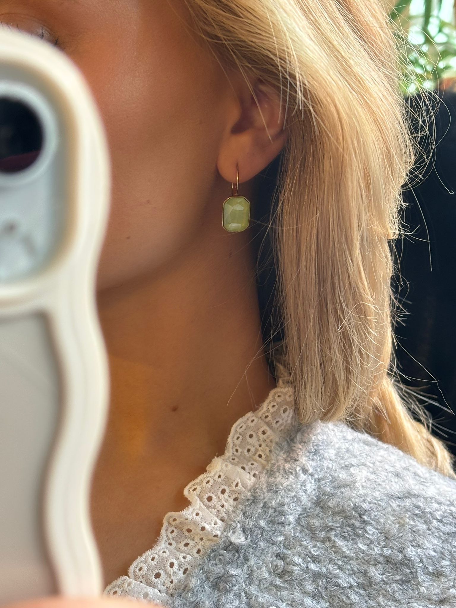 Närbild av en person med ljust hår som tar en spegelselfie och visar ett hängande örhänge i guld med rektangulär, ljusgrön sten. Personen bär grå stickad tröja och vit spetskrage i dagsljus.