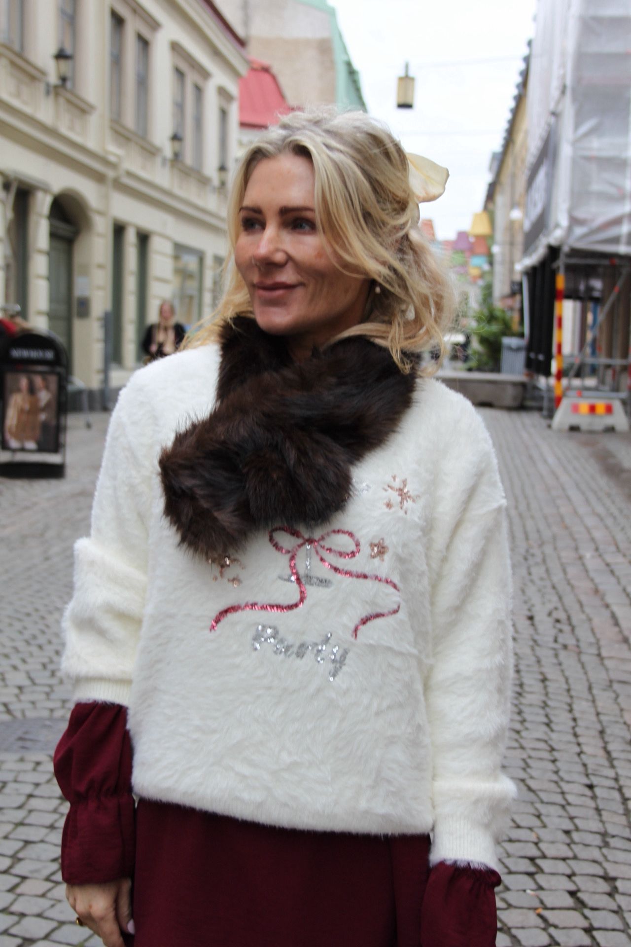 Leende blond kvinna står på en kullerstensgata och tittar åt sidan. Hon bär vit, fluffig tröja med mörk pälskrage och glittrig rosett samt texten ”Merry”, med stadshusfasader och byggställning i bakgrunden.
