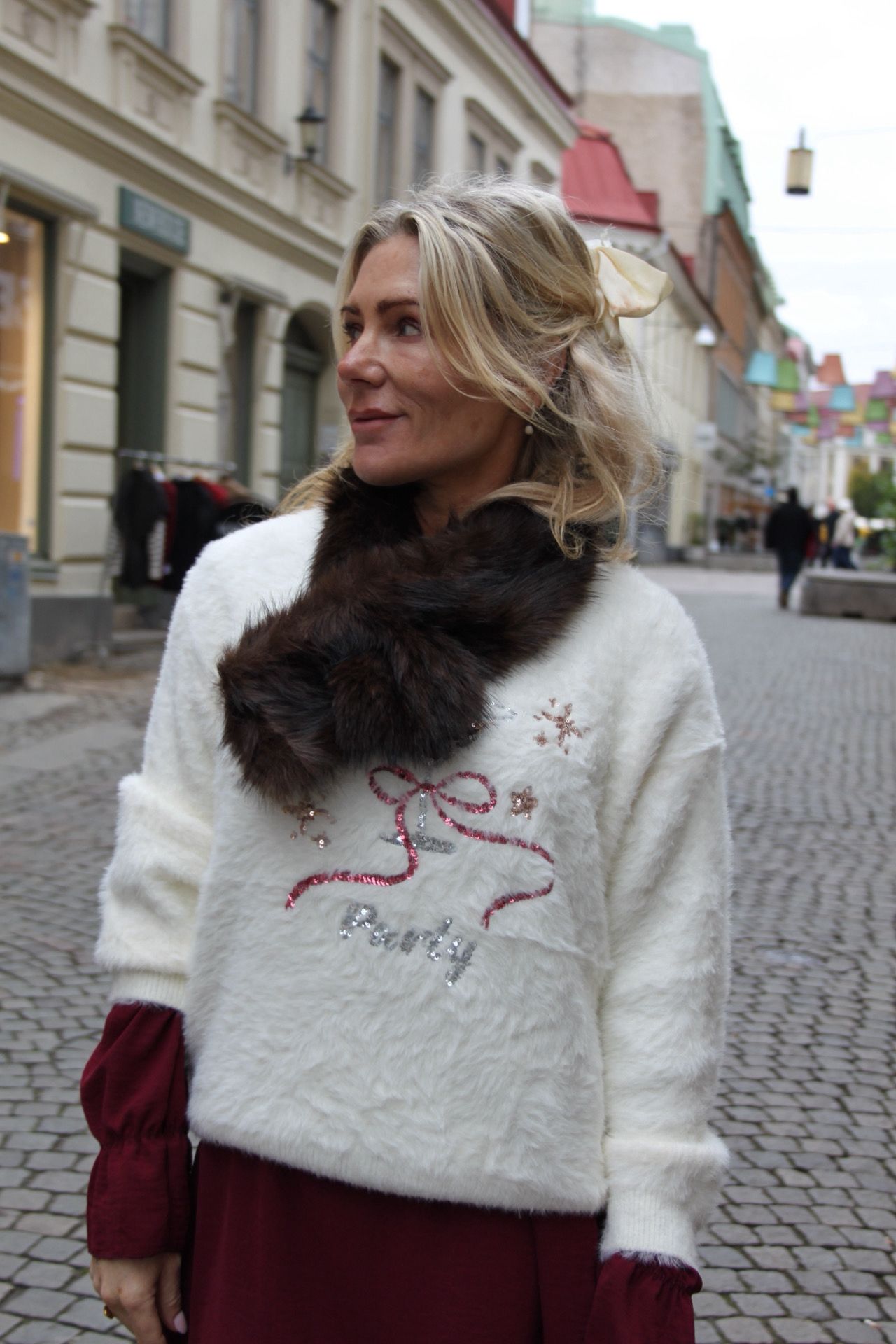 Kvinna med blont hår och ljus rosett tittar åt sidan på en kullerstensgata. Hon bär vit, lurvig tröja med paljettbroderi och texten ”Lovely” samt en mörk pälskrage, med stadshusfasader i bakgrunden.