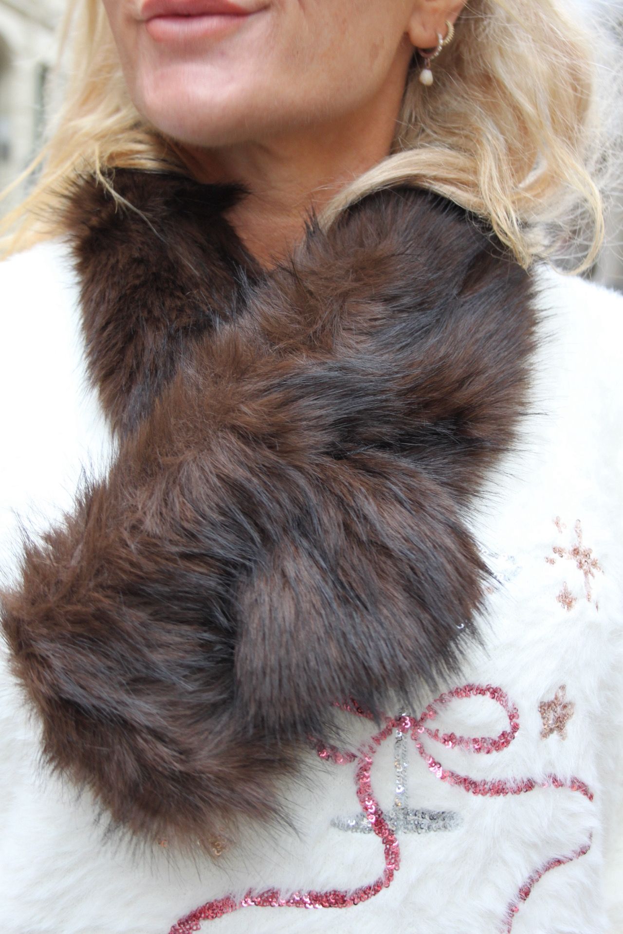 Närbild av en kvinna med blont hår som bär en stor mörkbrun pälskrage över en vit tröja. Hon står stilla och tittar bort från kameran; en liten örhänge syns och tröjan har glittrigt mönster.