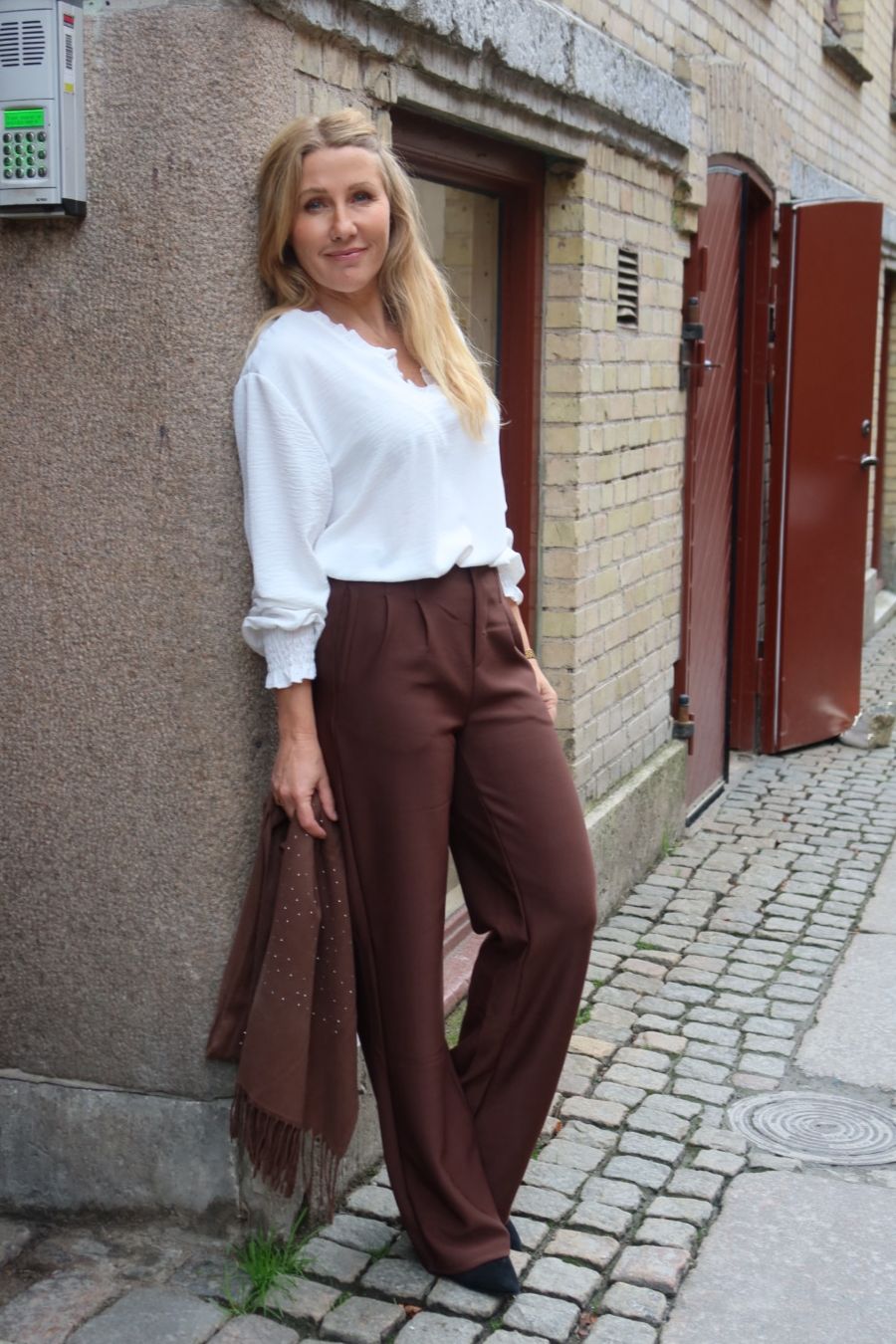 Blond kvinna lutar sig mot en vägg och ler mot kameran, klädd i vit blus och bruna, vida byxor. Hon håller en brun sjal med fransar i en smal kullerstensgränd med tegelvägg och röda dörrar.