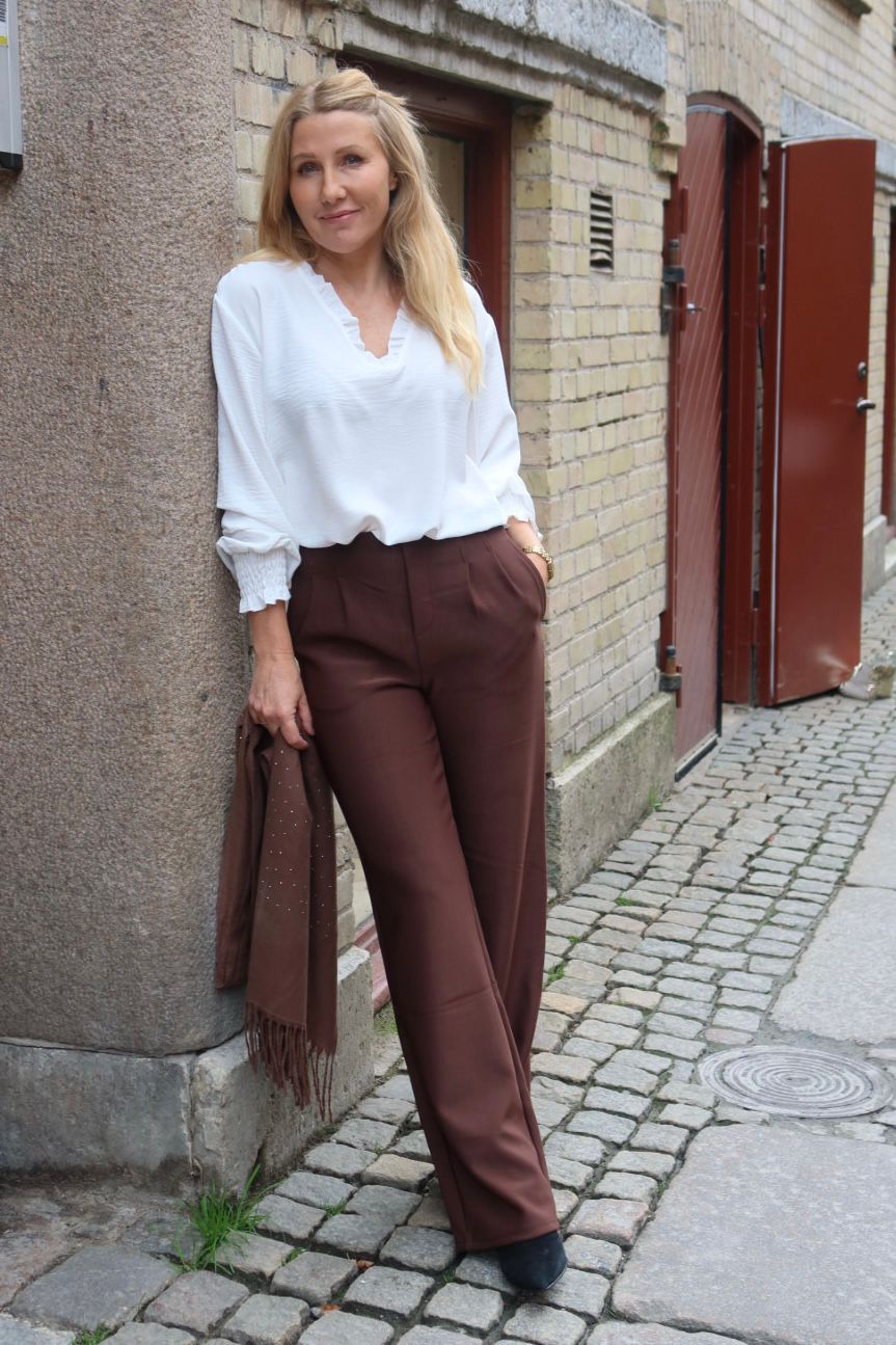 Blond kvinna lutar sig mot en husvägg och tittar mot kameran, med ena handen i fickan och en brun sjal i den andra. Hon bär vit blus och bruna byxor på en kullerstensgata.