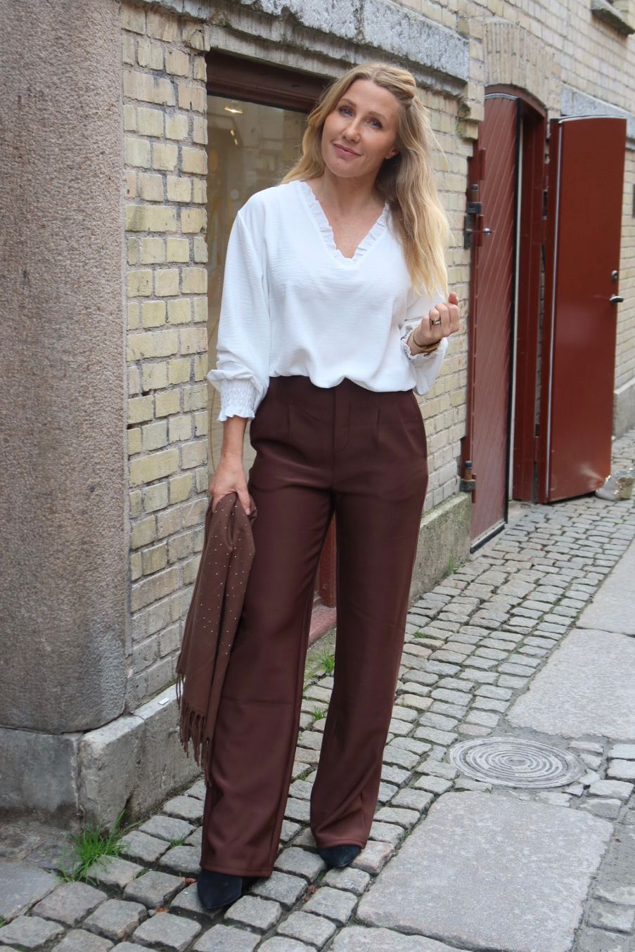 Blond kvinna står på en kullerstensgata vid en ljus tegelvägg och tittar mot kameran. Hon bär vit blus och bruna vida byxor, håller en brun sjal i handen och har röda portar i bakgrunden.