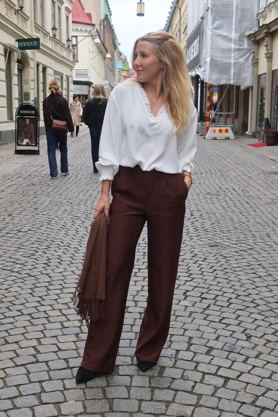 Kvinna med långt blont hår står på en kullerstensgata och tittar åt sidan, med ena handen i fickan. Hon bär vit blus, bruna vida byxor och håller en brun sjal; butiksskylten “NEWHOUSE” syns i bakgrunden.
