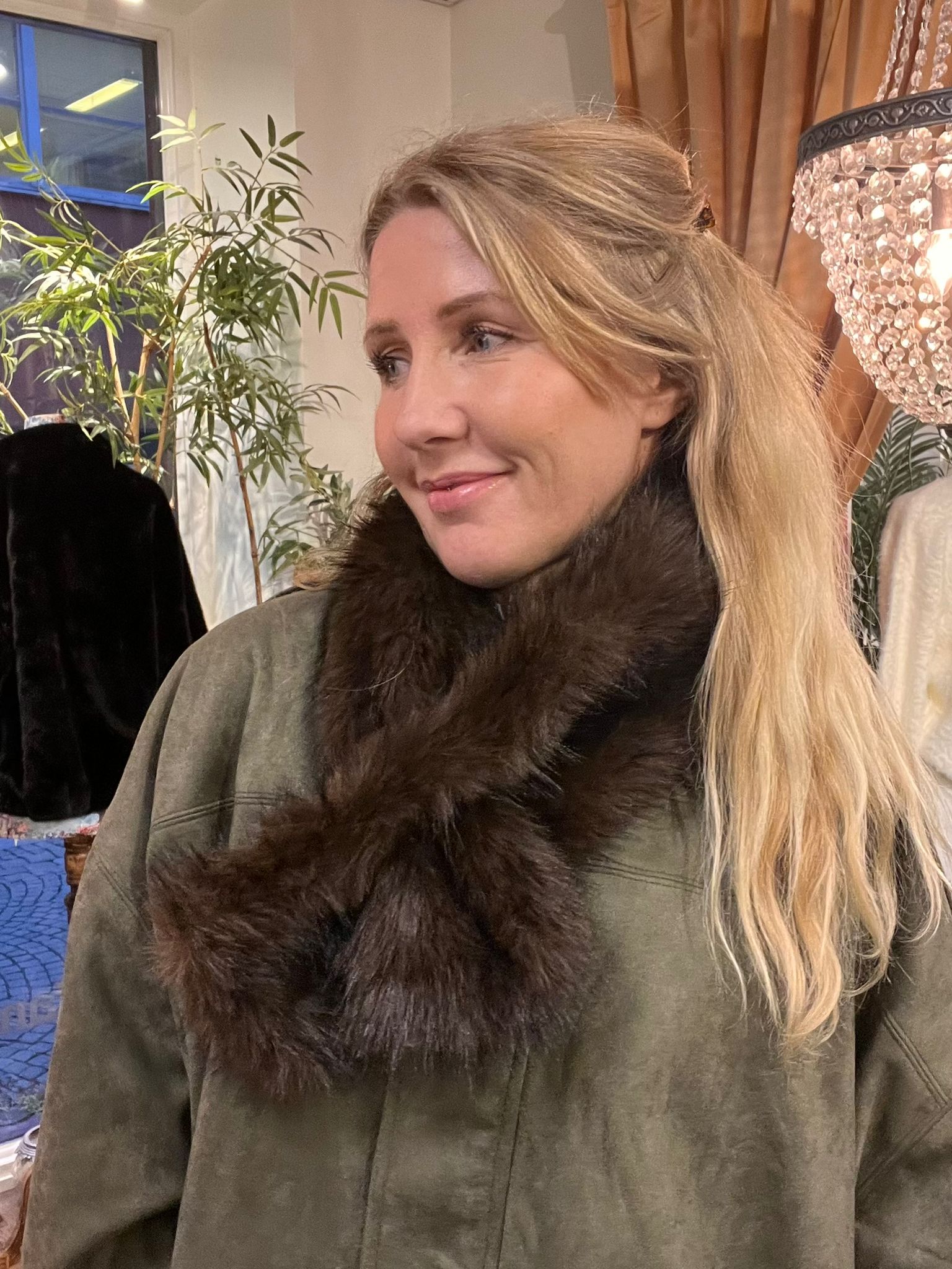 Blond kvinna med en mörk pälskrage över en olivgrön topp tittar åt sidan och ler svagt. Hon sitter inomhus nära en kristallampa och en stor grön växt, med gardiner i bakgrunden.