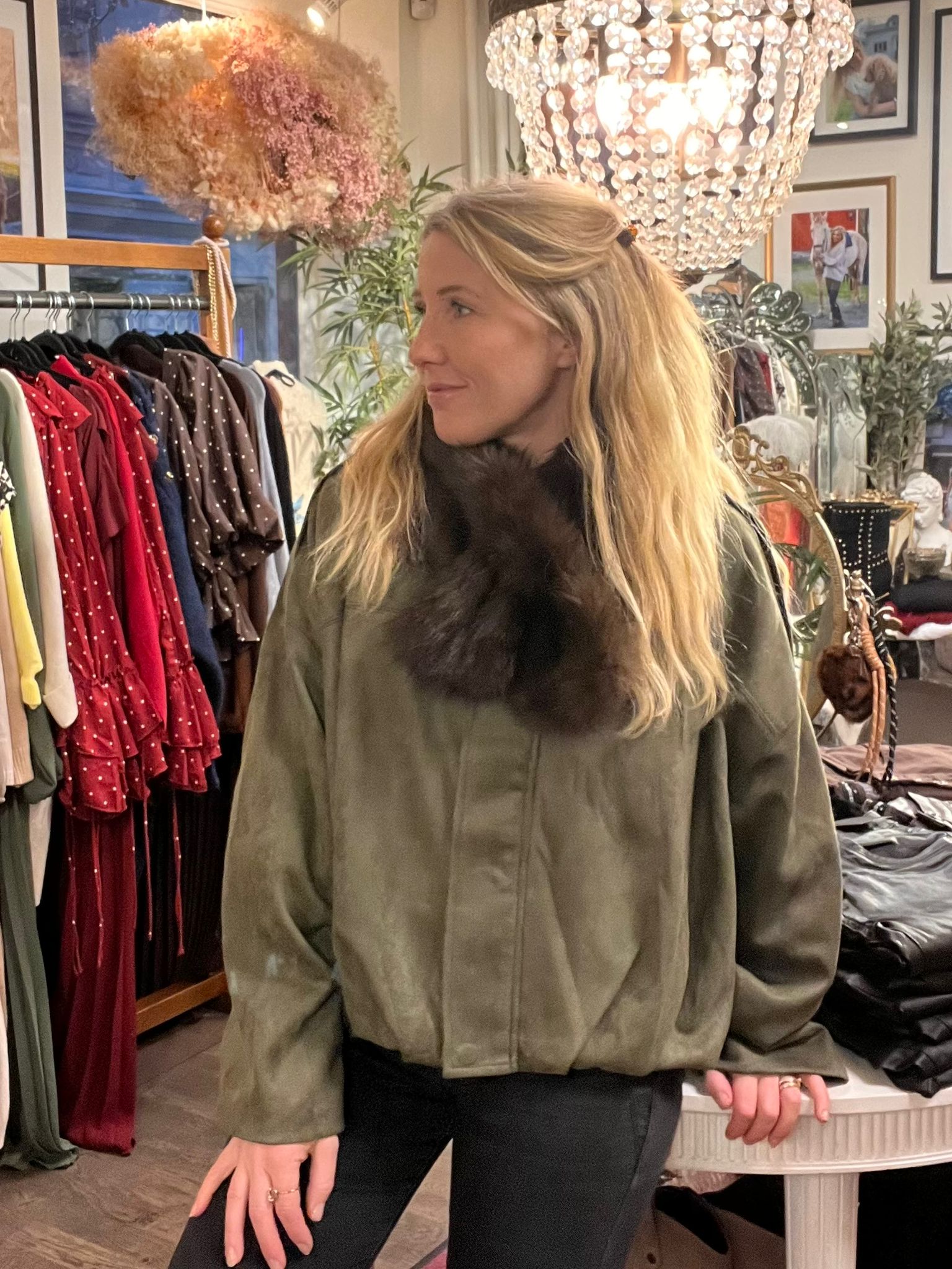 Blond person står inomhus och tittar åt sidan, klädd i olivgrön jacka med stor mörk pälskrage. I bakgrunden syns en klädbutik med klädställning, kristallkrona, växter och inramade foton på väggen.