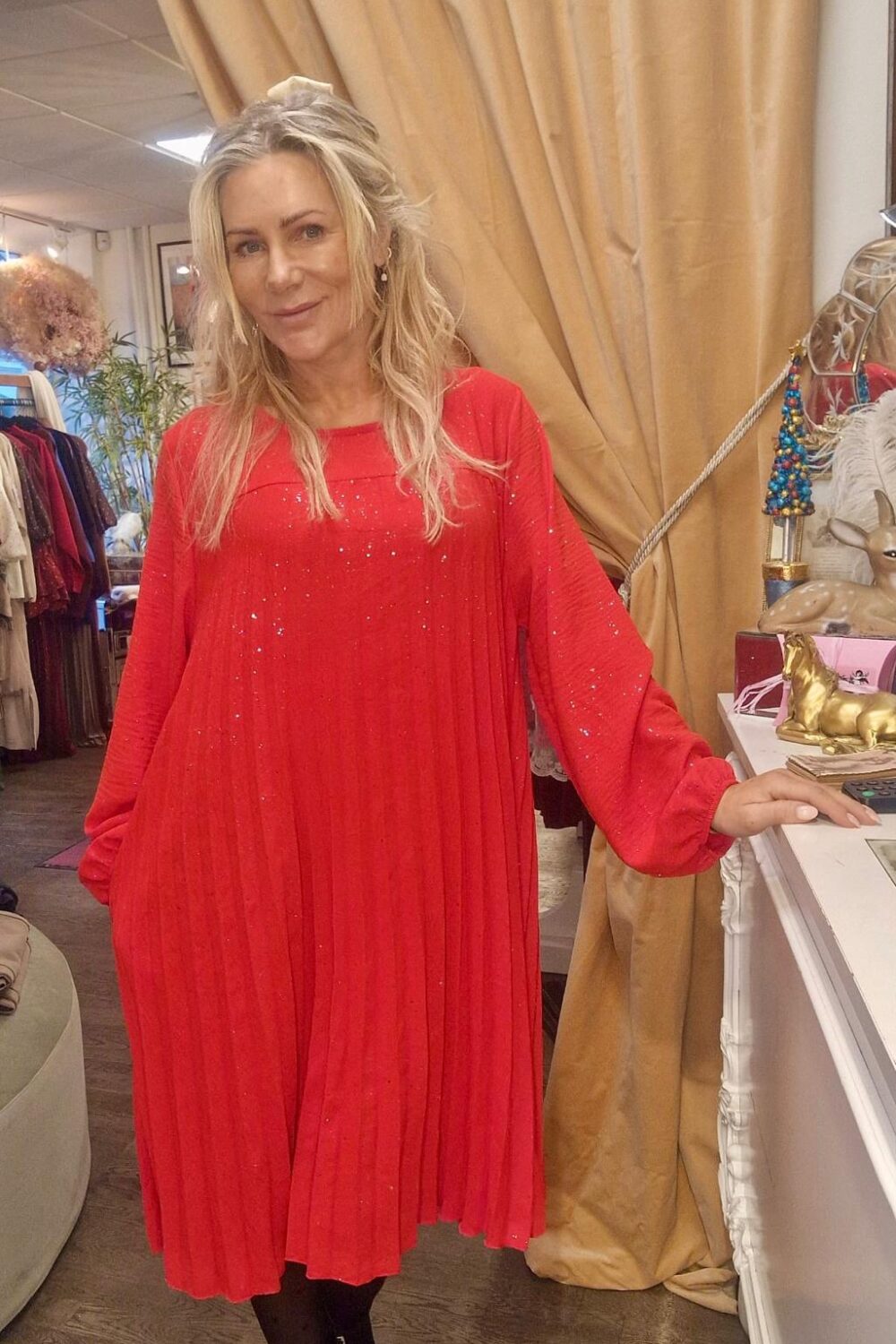 Kvinna i röd, glittrande och plisserad klänning står och ler i en butik, med ena handen mot en vit disk. I bakgrunden syns klädställningar, en beige gardin och dekorföremål.