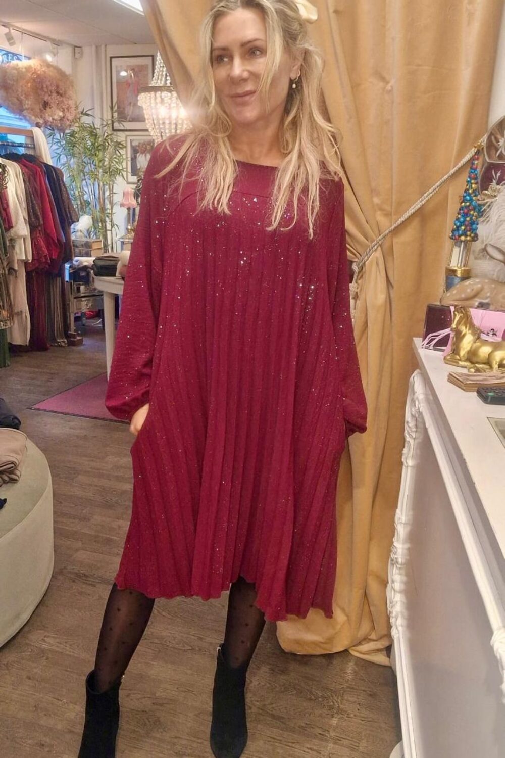Kvinna i en vinröd, glittrig och plisserad klänning står med händerna i fickorna och tittar åt sidan. Hon bär svarta strumpbyxor och ankelboots, i en klädbutik med klädställningar och beige draperi.