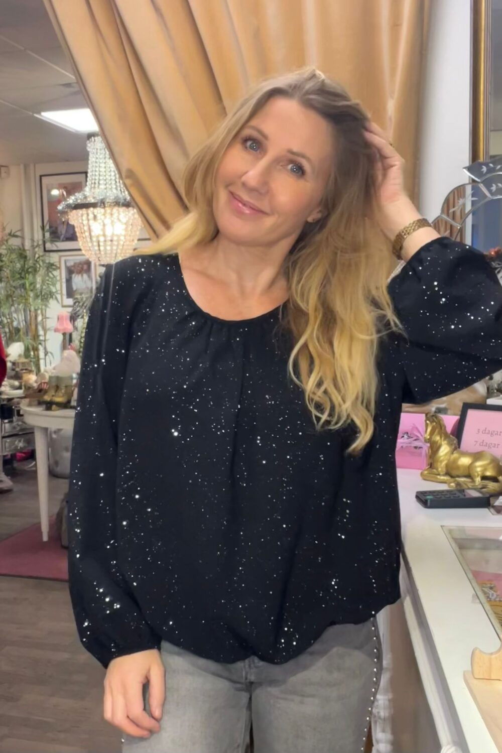 Leende kvinna med långt blont hår lutar sig mot en disk och lyfter handen till håret. Hon bär en svart glittrig blus och grå jeans, i en butiksmiljö med kristallkrona, draperier och smyckesställ.