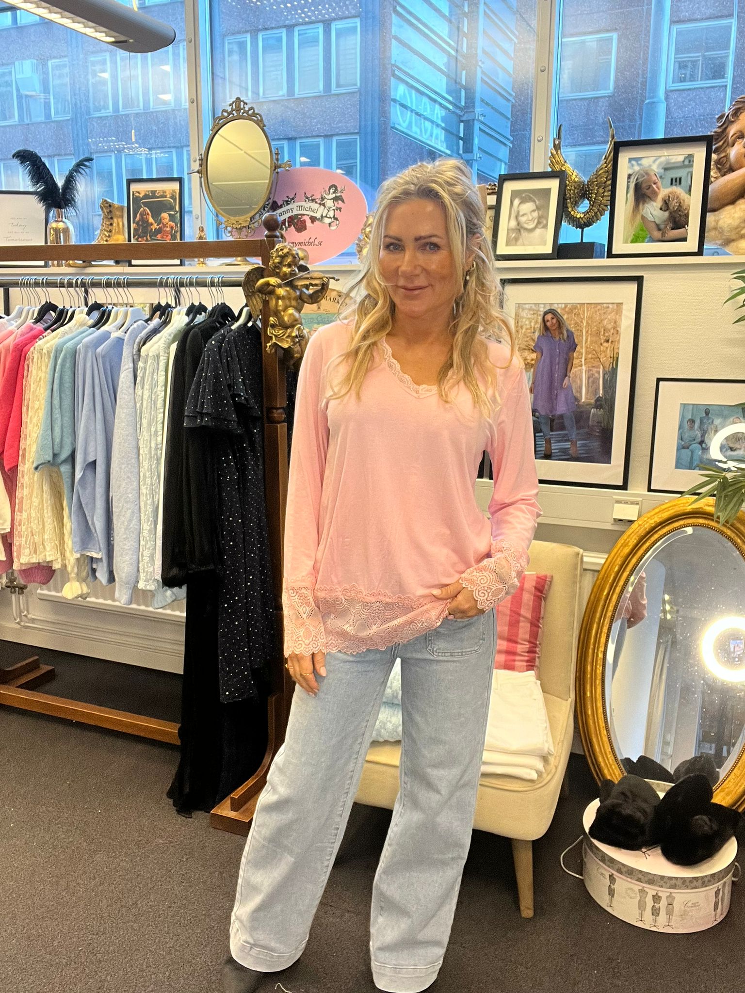 Kvinna står och ler i en klädbutik, iklädd ljusrosa blus med spets och ljusa jeans. Bakom henne syns klädställning med plagg, speglar, änglafigur, inramade foton och texten “Jenny Michel”.