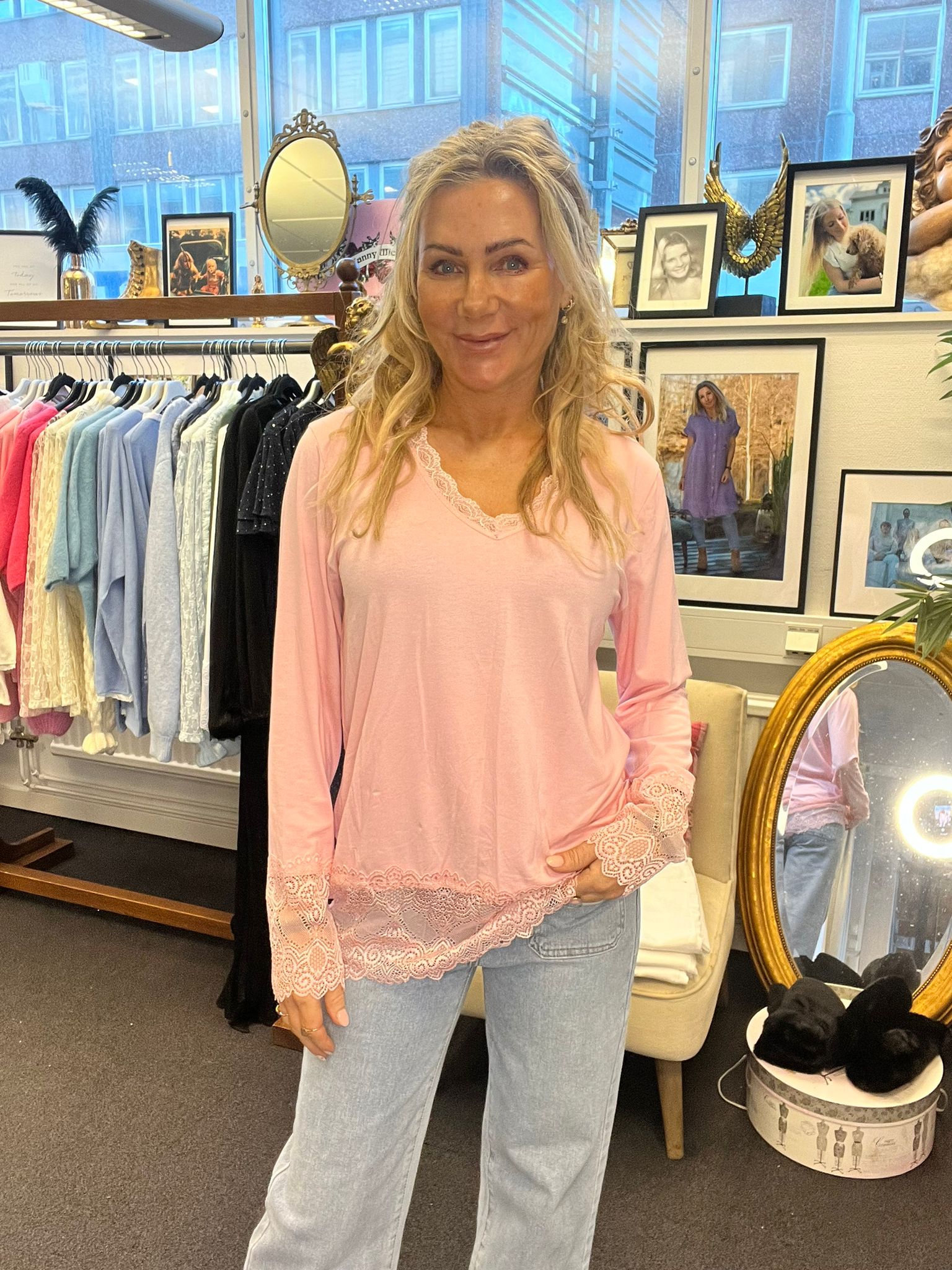 Blond kvinna står och ler mot kameran i en klädbutik, iklädd rosa topp med spetsdetaljer och ljusa jeans. Bakom henne syns klädställning med pastellplagg, speglar och inramade foton på en hylla.