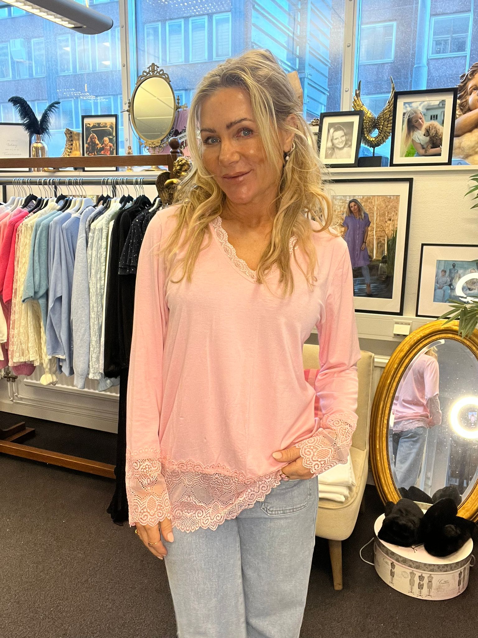 Kvinna med långt blont hår står och ler i en butiksmiljö, iklädd ljusrosa topp med spetskant och ljusa jeans. Bakom henne syns klädställning med plagg samt speglar och inramade fotografier på en hylla.