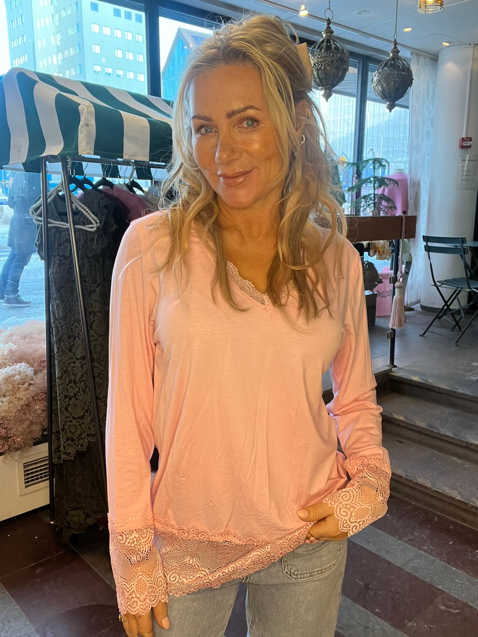 Blond kvinna ler mot kameran och håller händerna framför magen, klädd i rosa långärmad topp med spetsdetaljer och jeans. Hon står inomhus nära ett klädställ och stora fönster med stadshus i bakgrunden.