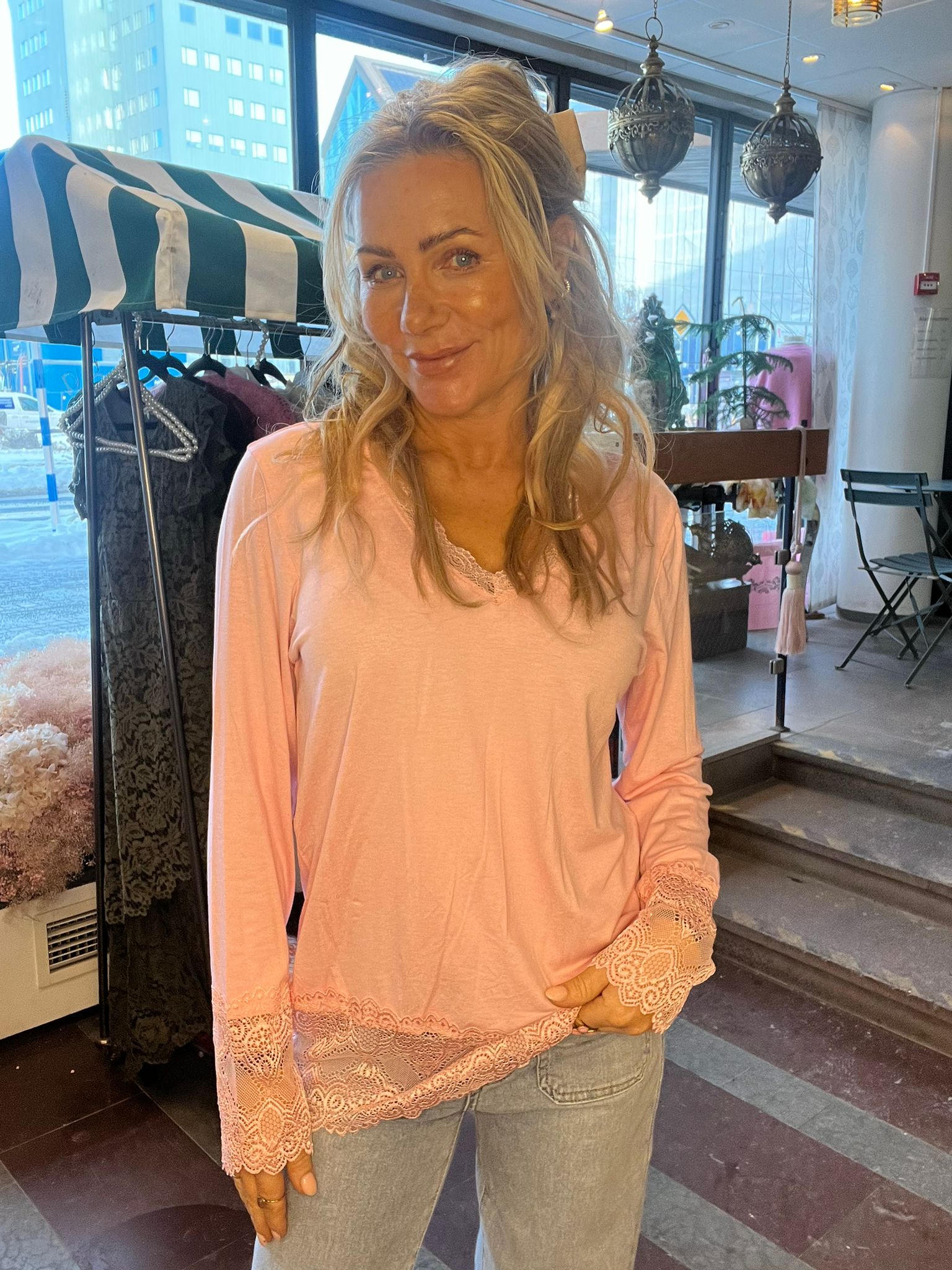 Blond kvinna står och ler mot kameran i en butiksmiljö, klädd i ljusrosa topp med spetskant och ljusa jeans. Bakom henne syns klädställning, randig markis, hänglampor och stora fönster mot stadsmiljö.