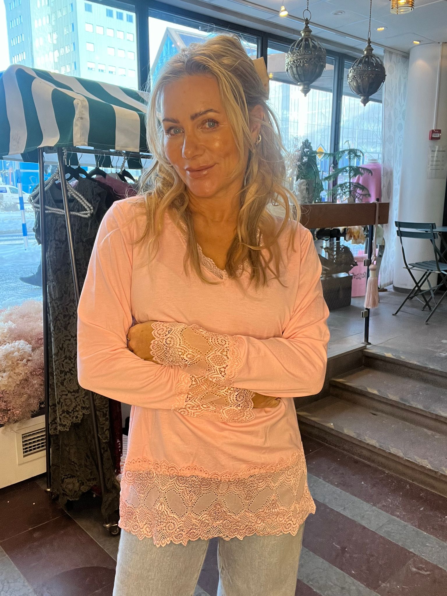 Blond kvinna står med armarna i kors och ler mot kameran, klädd i ljusrosa topp med spetsdetaljer och ljusa byxor. Hon står i en butiksmiljö med klädställning, stora fönster och hänglampor.