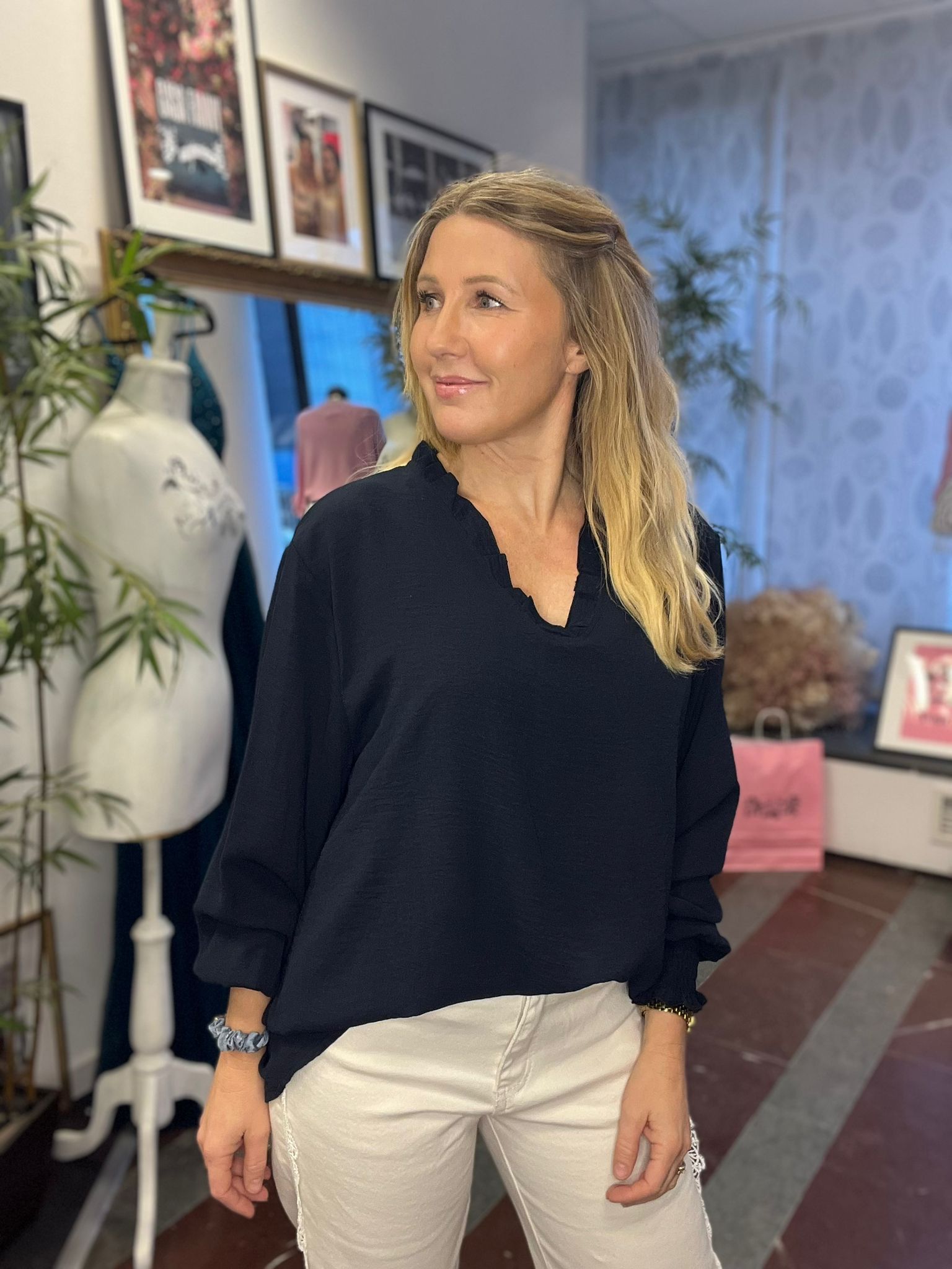 Blond kvinna i svart blus och vita byxor poserar och ler lätt medan hon tittar åt sidan. Hon står inomhus i en butik eller studio med provdocka, växt och inramade bilder i bakgrunden.