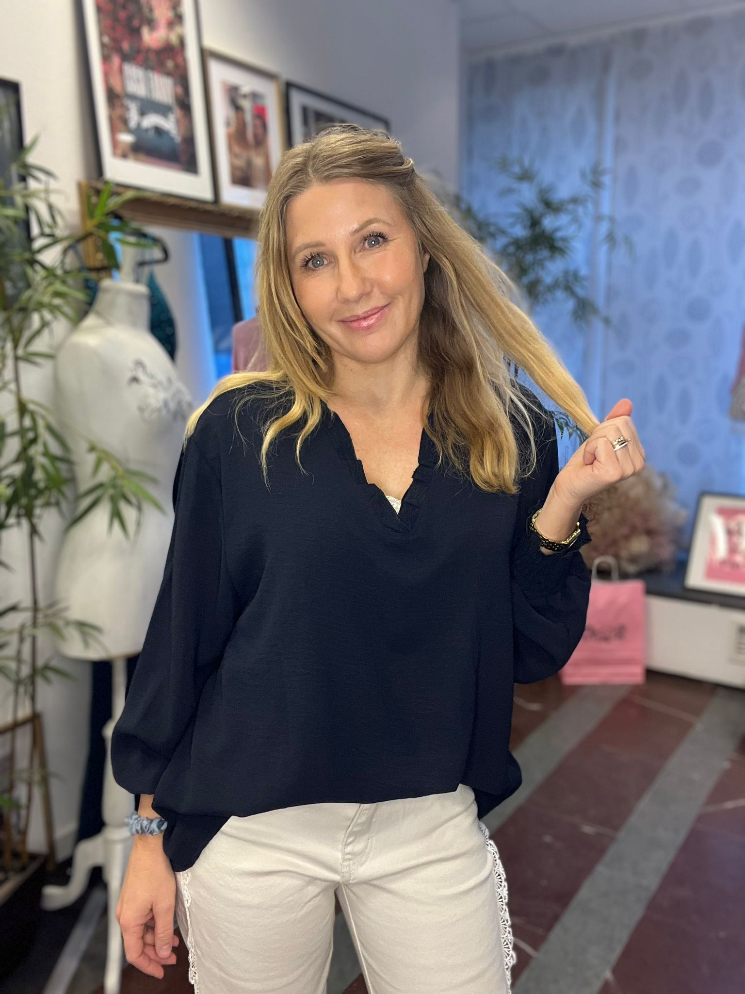 Leende kvinna med långt blont hår håller upp en hårslinga och poserar i en mörk blus och ljusa byxor. Hon står inomhus i ett rum med provdocka, växter och inramade bilder på väggen.