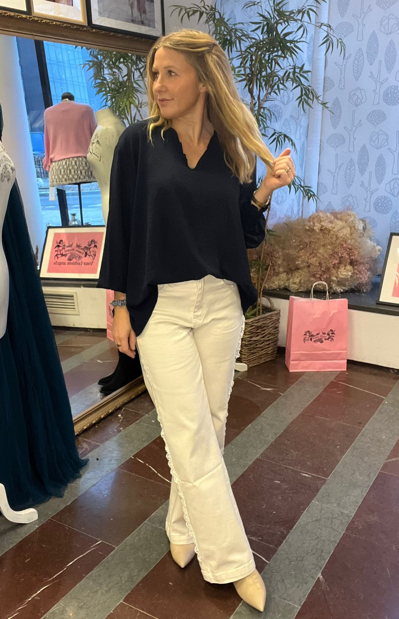 Blond kvinna i svart blus och vita byxor poserar och håller i en hårslinga medan hon tittar åt sidan i en butiksmiljö. I bakgrunden syns spegel, provdocka, växter och rosa påsar. Text: “Your fashion mates”.