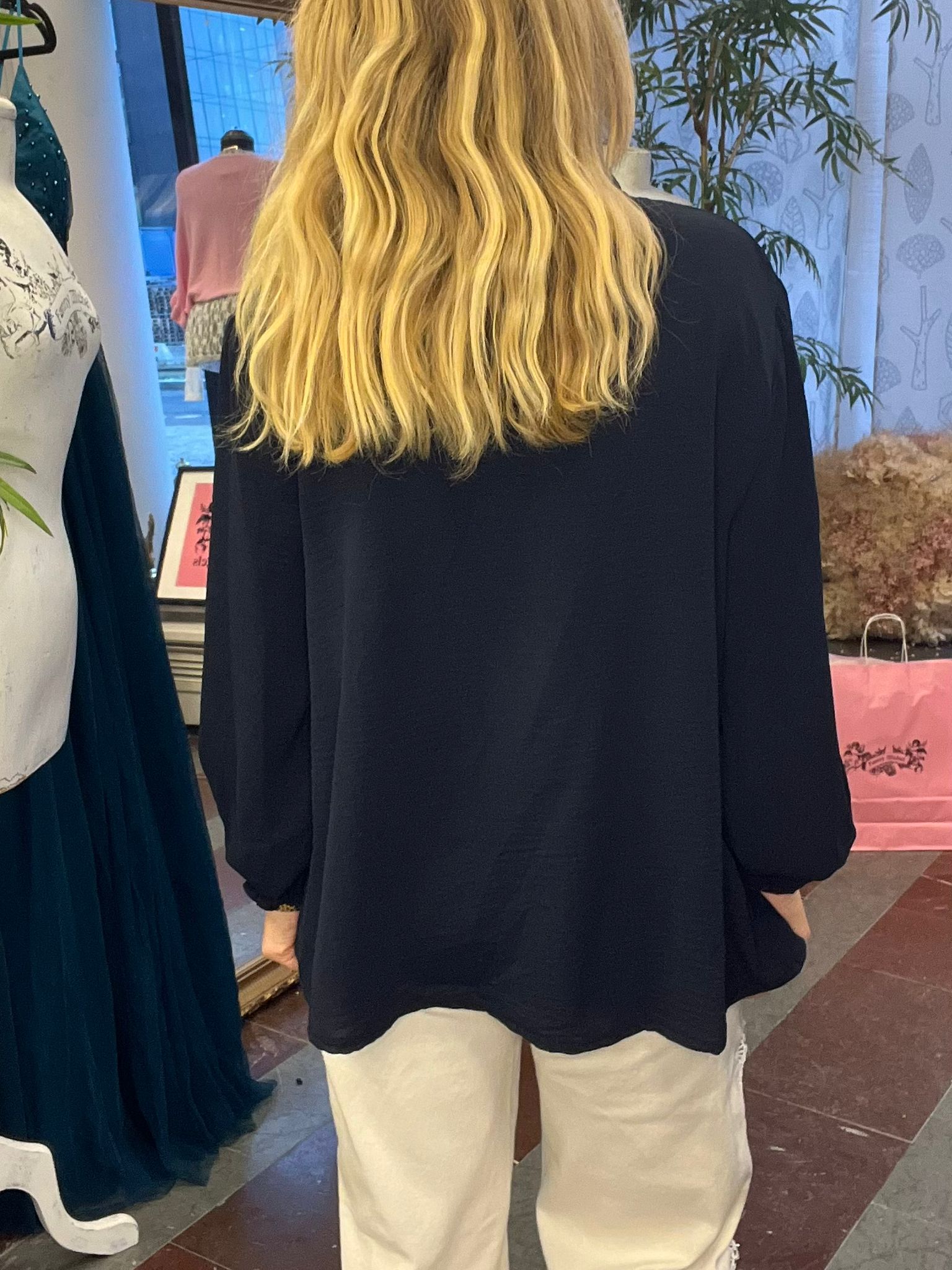 Person med långt, vågigt blont hår står med ryggen mot kameran och bär en mörk blazer och ljusa byxor, i en butiksmiljö med klänningar på skyltdockor, växter och rosa shoppingkasse i bakgrunden.