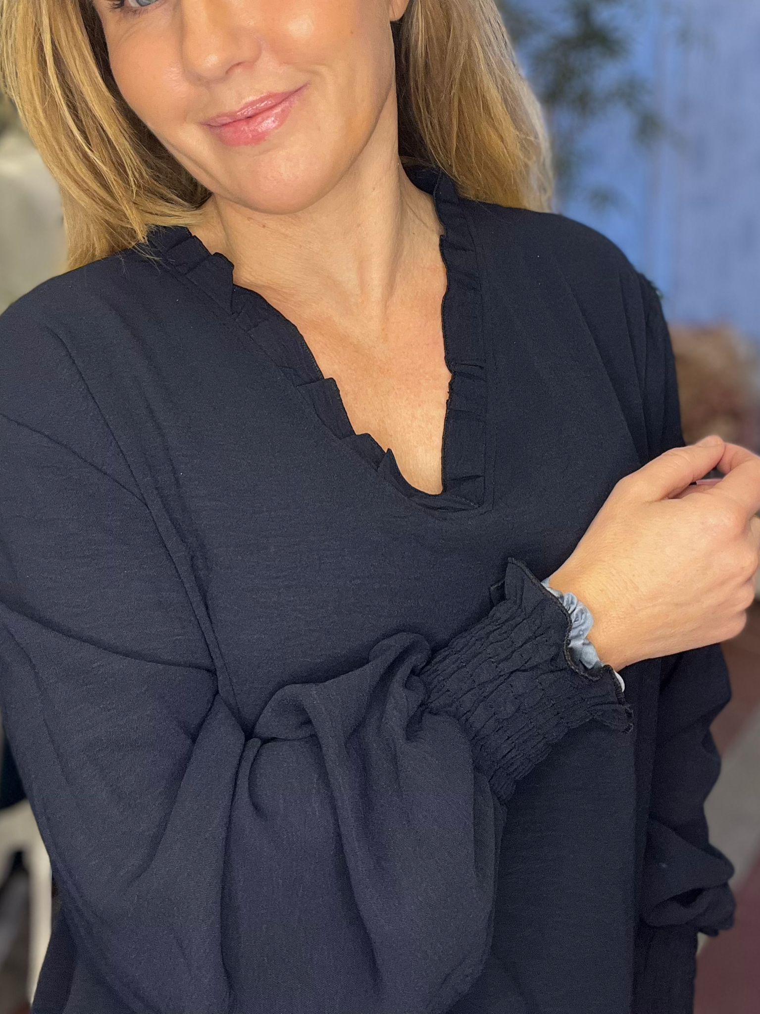 Närbild av en leende person med blondt hår som håller handen mot bröstet och visar en mörkblå blus med volangkrage och rynkade ärmslut med ljus scrunchie. Bakgrunden är mjukt suddig inomhus.