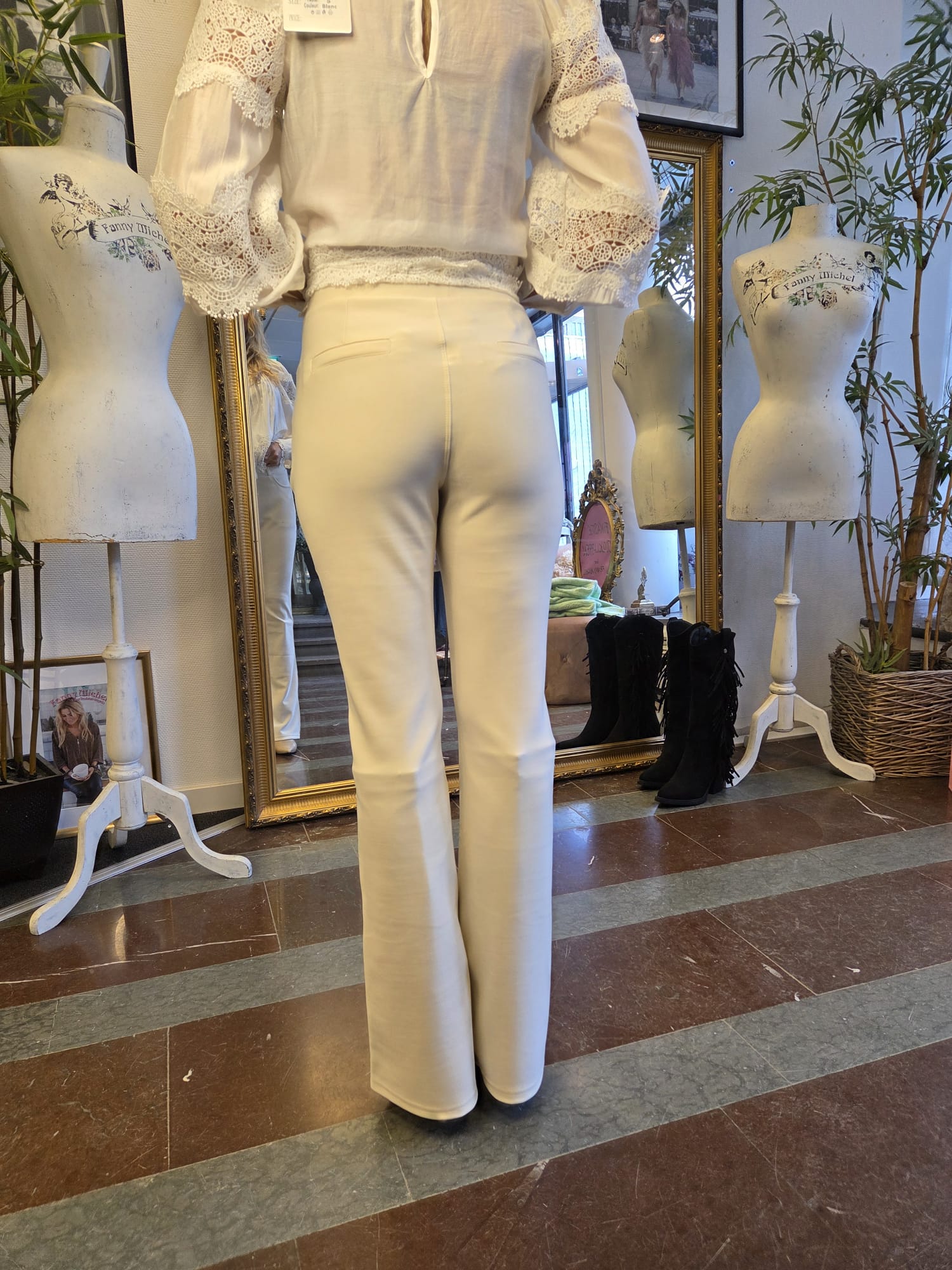 Person står med ryggen mot kameran och provar vita utsvängda byxor och en vit blus med spetsärmar framför en stor spegel i en butik, omgiven av provdockor med texten “Fanny Michel” och svarta fransstövlar.