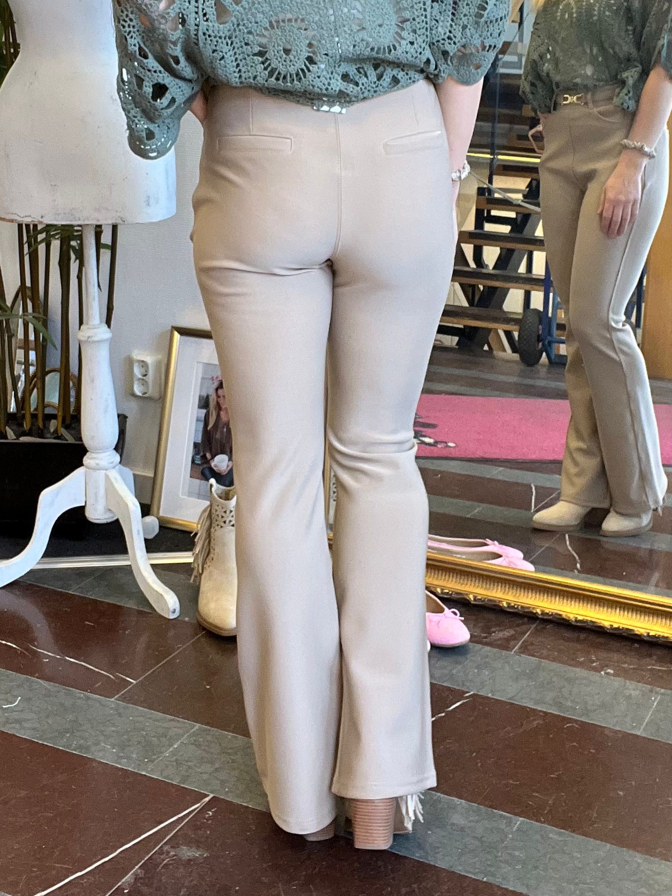 Person står med ryggen mot kameran och provar ljusbeige, utsvängda byxor med grön spetsblus. Spegeln visar sidovy och framsida. Miljön liknar en butik med provdocka, guldramsspegel och rosa skor på golvet.