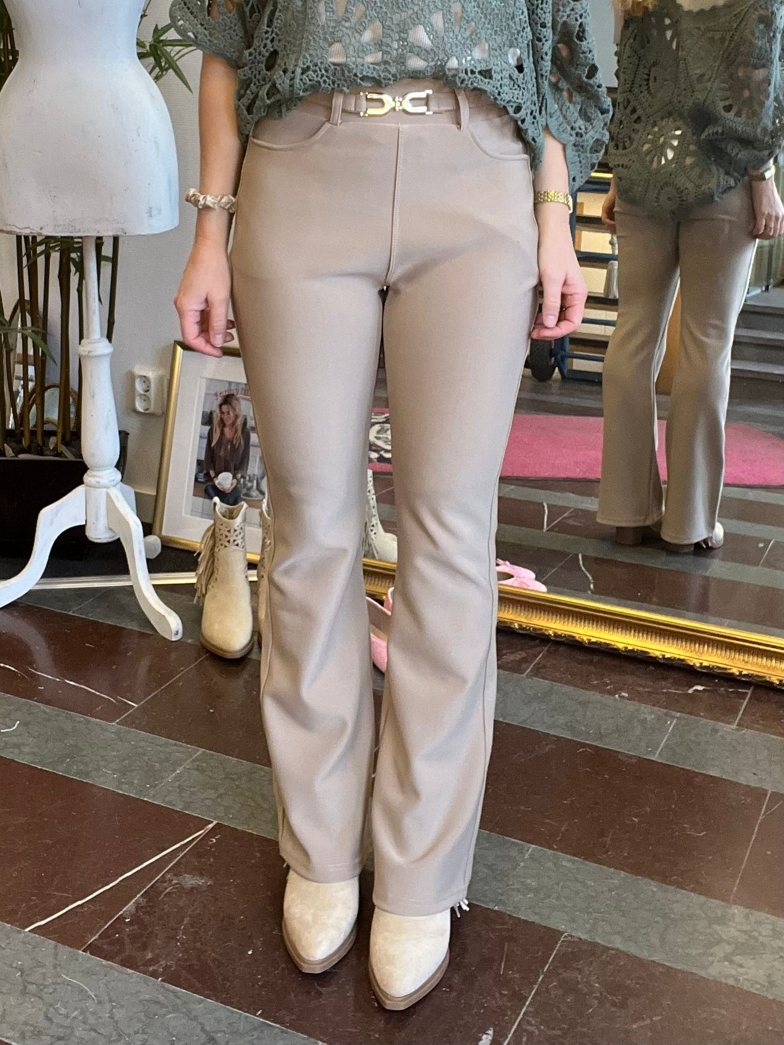 Person står i en butik och visar ljusbeige, lätt utsvängda byxor med bälte och en grön virkad topp. I spegeln syns samma outfit bakifrån; vita boots och ett provrumsområde i bakgrunden.