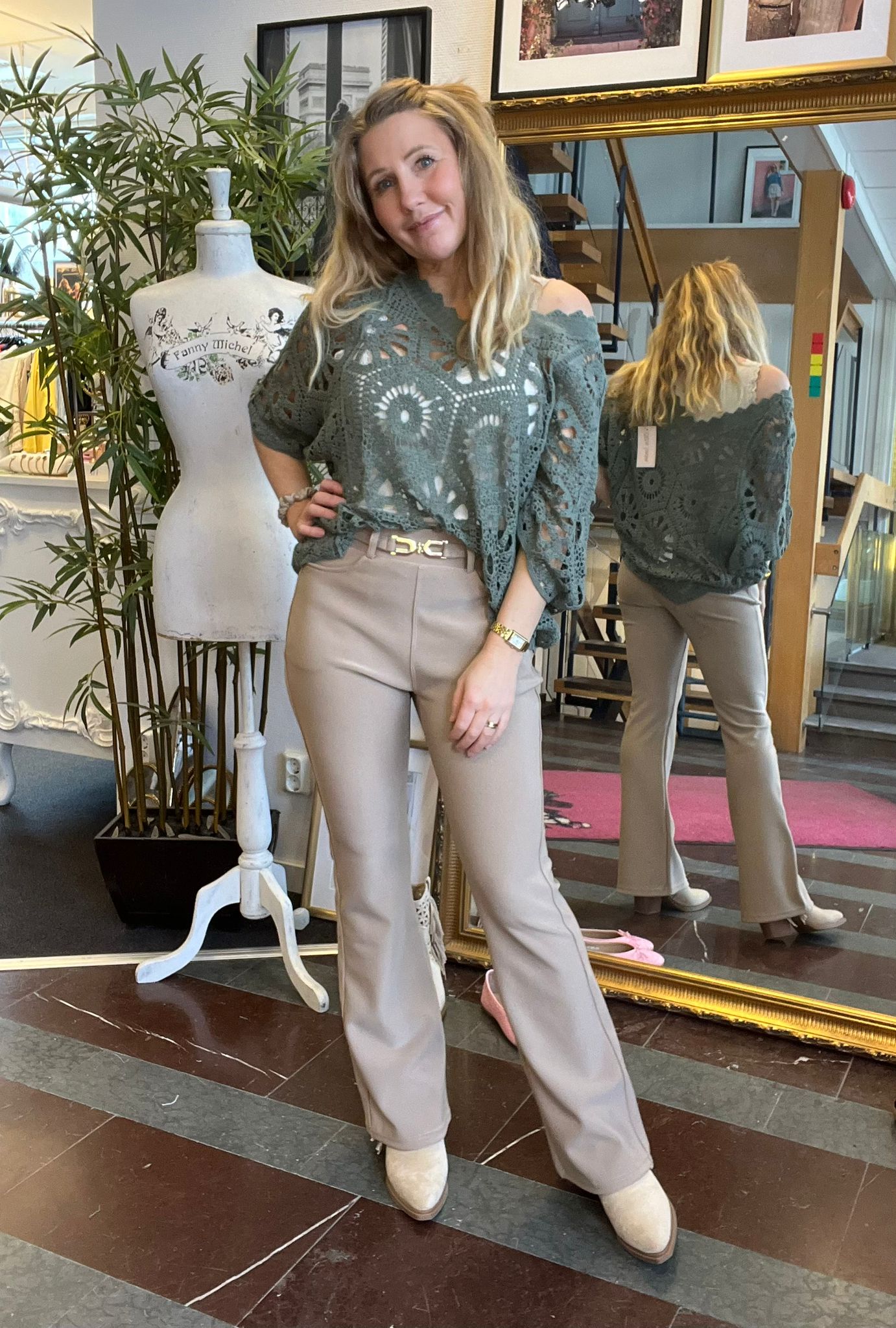 Kvinna poserar med handen i midjan i en klädbutik, klädd i grön virkad topp och beige byxor, med sin rygg synlig i en stor spegel. Bredvid står en provdocka med texten “Fanny Michel”.