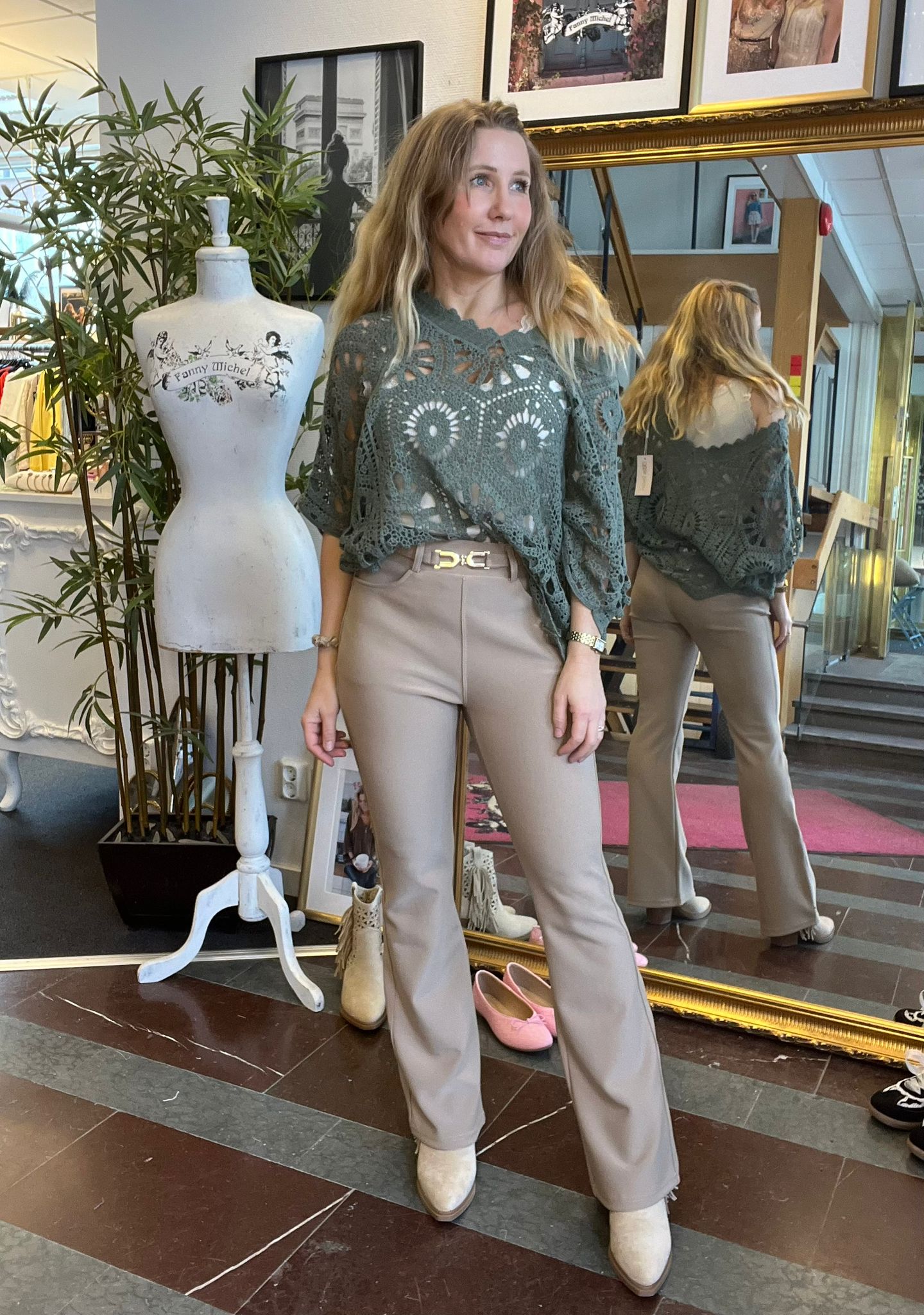 Kvinna står och poserar i en klädbutik framför en stor guldspegel, klädd i grön virkad topp, beige byxor och ljusa boots. Bredvid syns en provdocka med texten “Fanny Michel” och ett par rosa skor på golvet.
