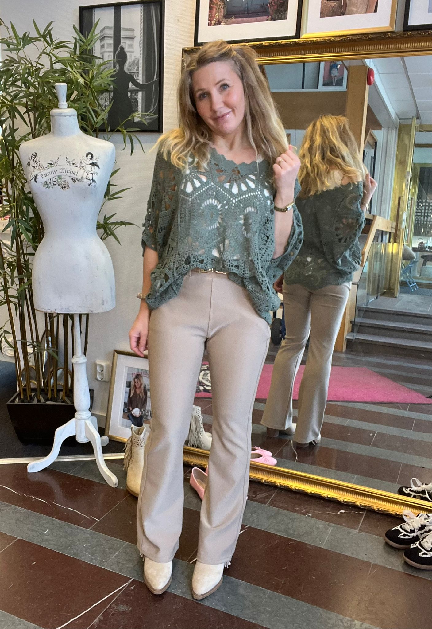 Kvinna poserar och visar en outfit med grön virkad topp, beige byxor och ljusa boots i en inredningsbutik. Hon står framför en stor guldspegel som speglar ryggen; intill syns provdocka med texten ”Fanny Michel”.