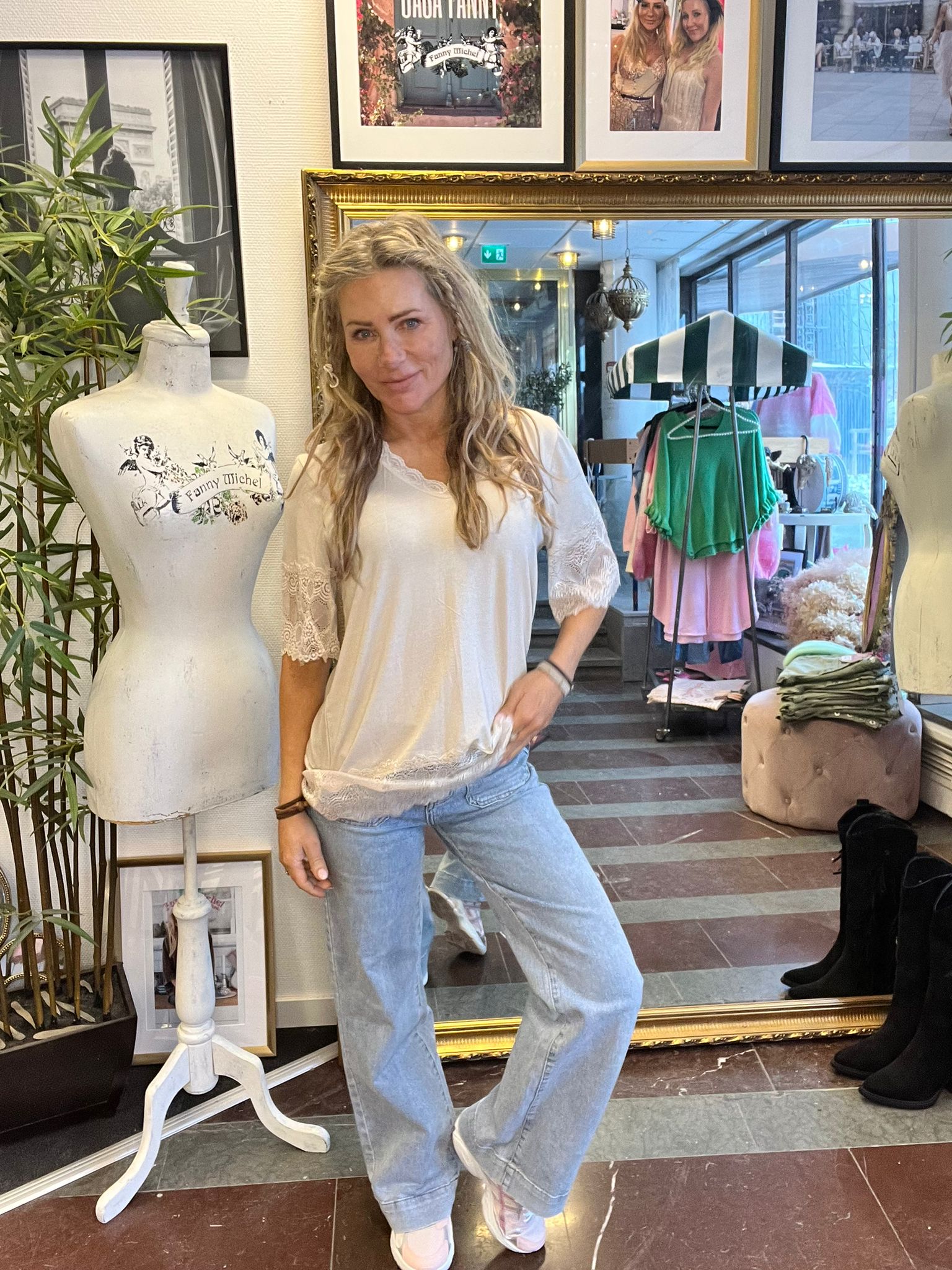 Kvinna i vit blus och jeans poserar och håller i blusens fåll inne i en klädbutik, bredvid en provdocka med texten ”Fanny Michel”. I bakgrunden syns klädställning med färgglada plagg och inramade foton.