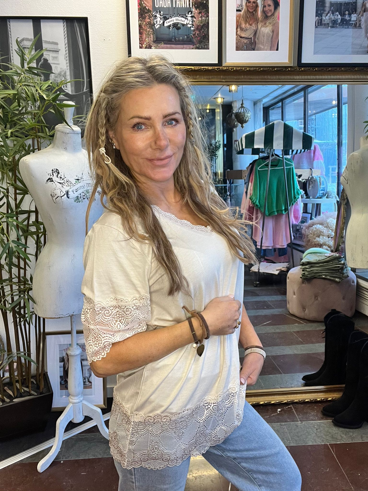 Leende kvinna poserar i vit tunika med spets och jeans bredvid en provdocka i en klädbutik. Bakom syns spegel, klädställning under randigt parasoll och inramade foton; text: ”OHOH FANNY” och ”Fanny Michel”.