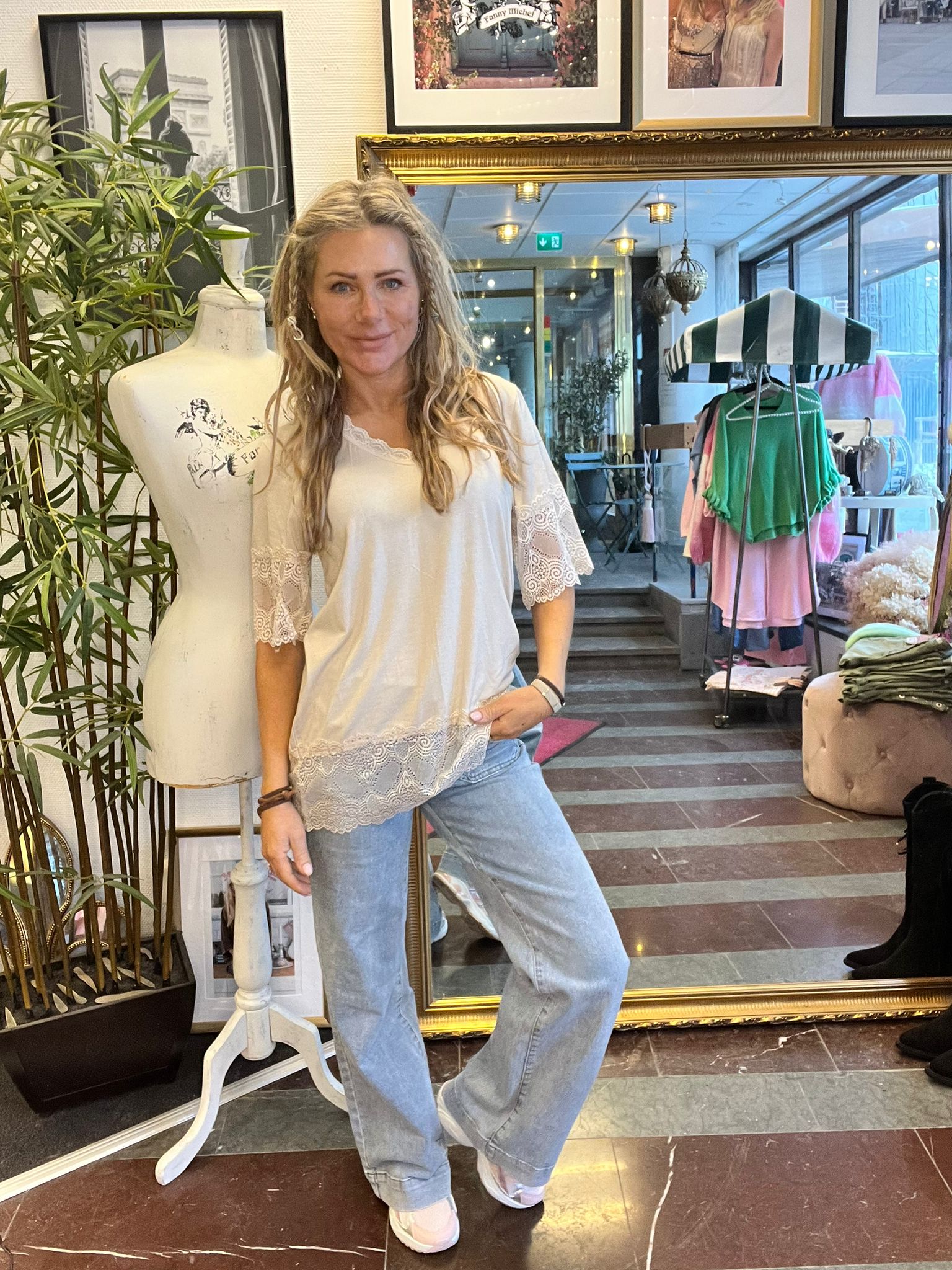 Kvinna i vit spetskantad blus och ljusa jeans poserar med handen i fickan bredvid en provdocka i en klädbutik. I bakgrunden syns klädställ med färgglada plagg, stor guldram och speglar. Text: “Fanny Michel”.
